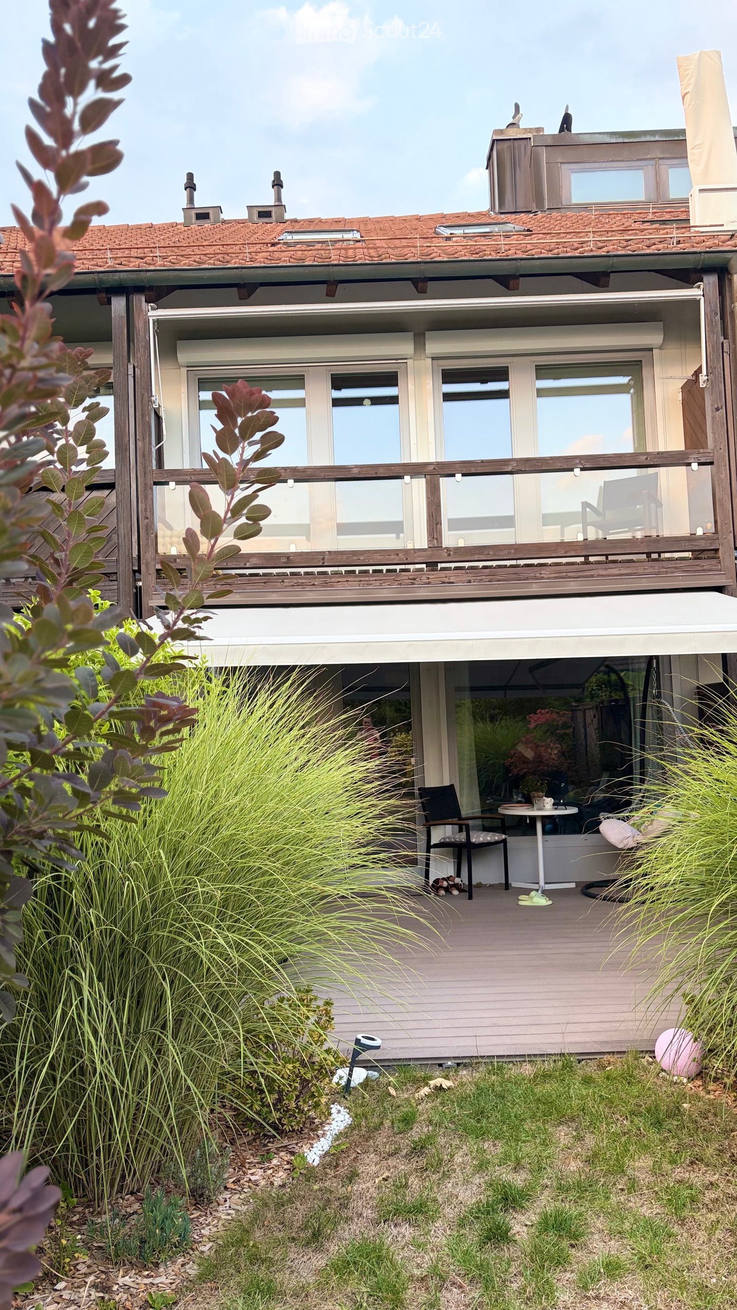 A modern two-story house with a wooden balcony and a well-maintained garden area featuring tall grasses, shrubs, and a small seating area with a table and chairs.