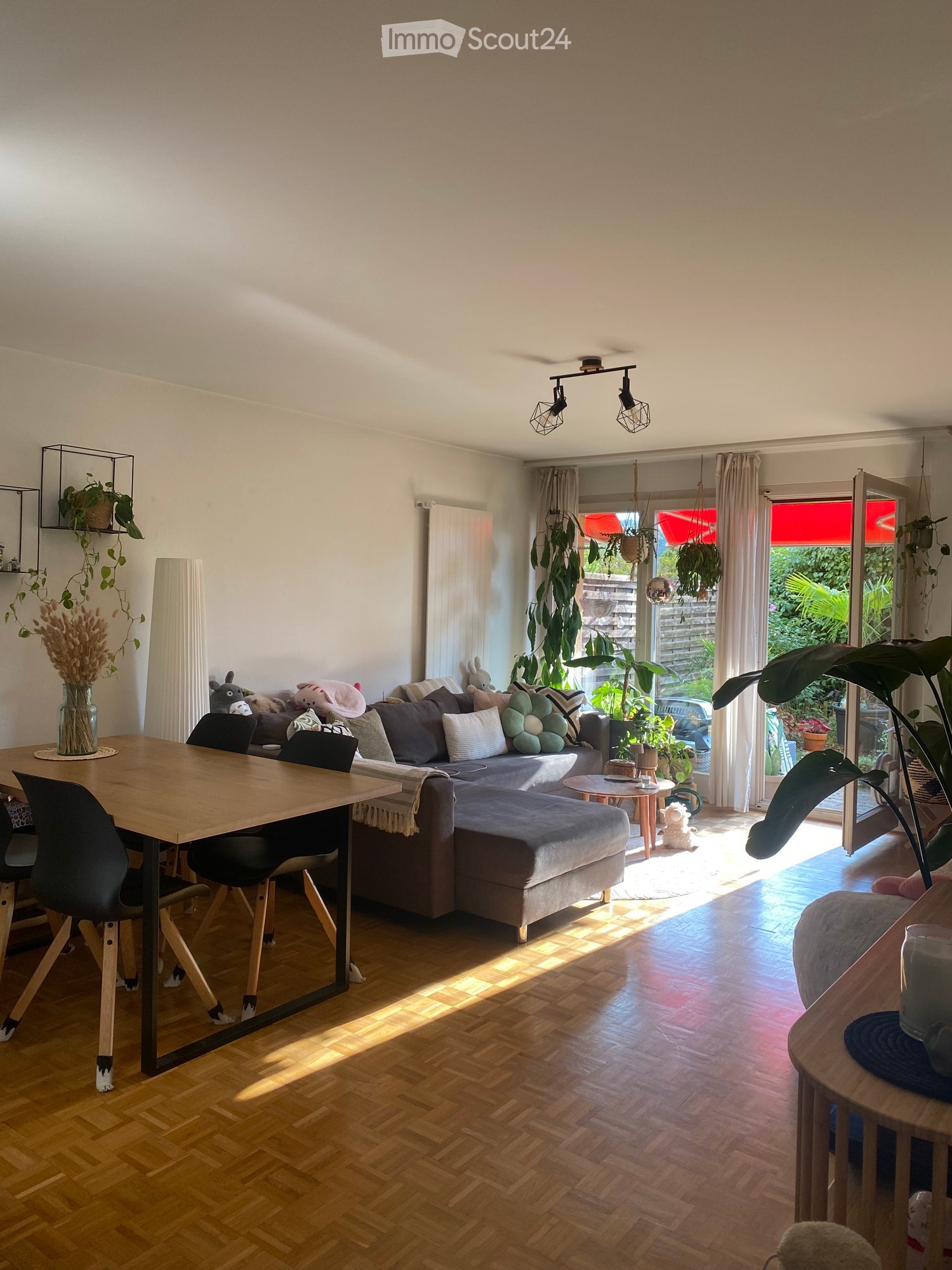 Bright living room with wooden flooring, large window with sheer curtains, and a sliding glass door leading to a garden. Features a grey sectional sofa with multiple cushions, a wooden coffee table, a dining table with chairs, and several potted plants.