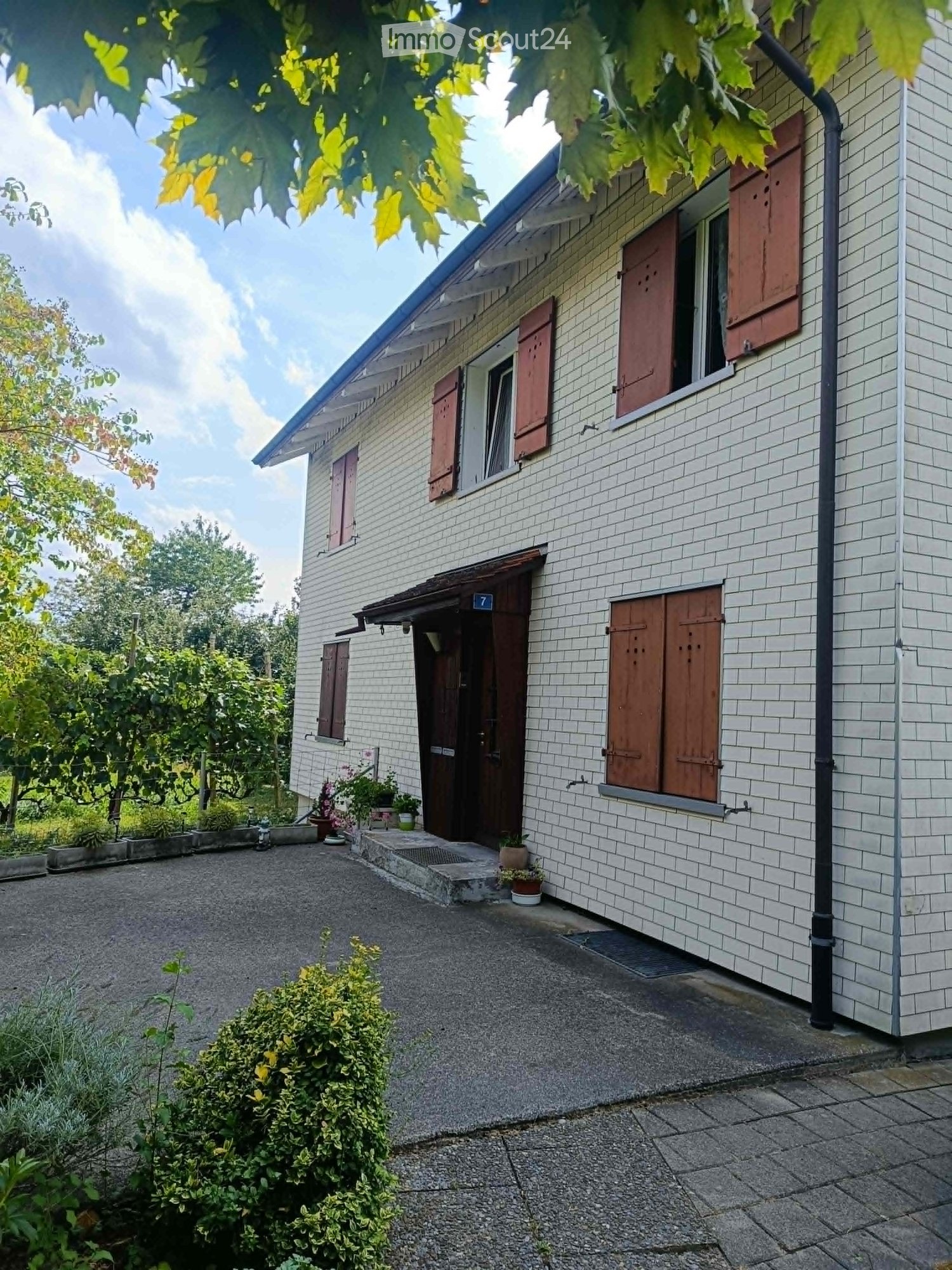 Two-story house with white brick exterior, red shutters, and a gravel driveway. The entrance features a small porch with potted plants and a small garden area with shrubs and trees.