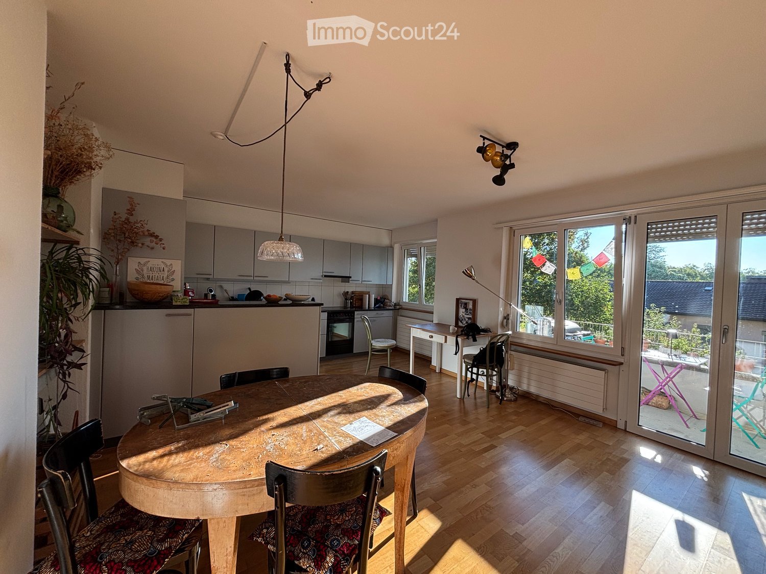 Bright, open-plan kitchen and dining area with wooden flooring, large windows, and modern grey cabinets. Features a round wooden dining table with chairs, a small kitchen island, and various appliances.