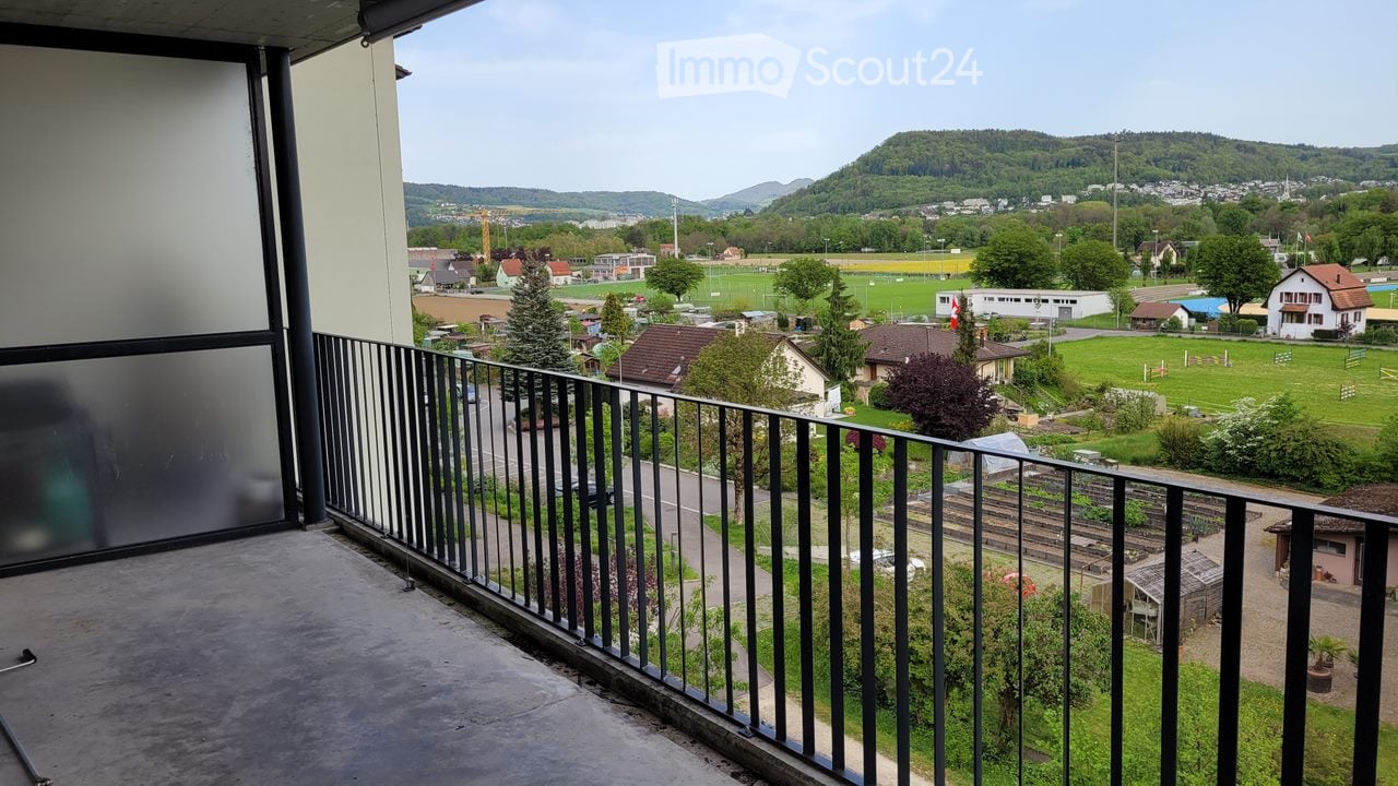 Balcony with metal railing overlooking a town and green fields, with distant hills and scattered houses.