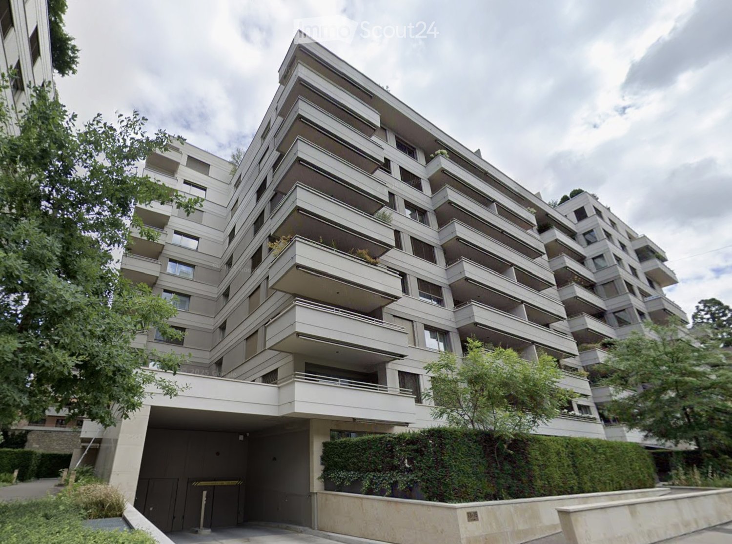 Modern multi-story residential building with balconies and a garage entrance, surrounded by greenery.