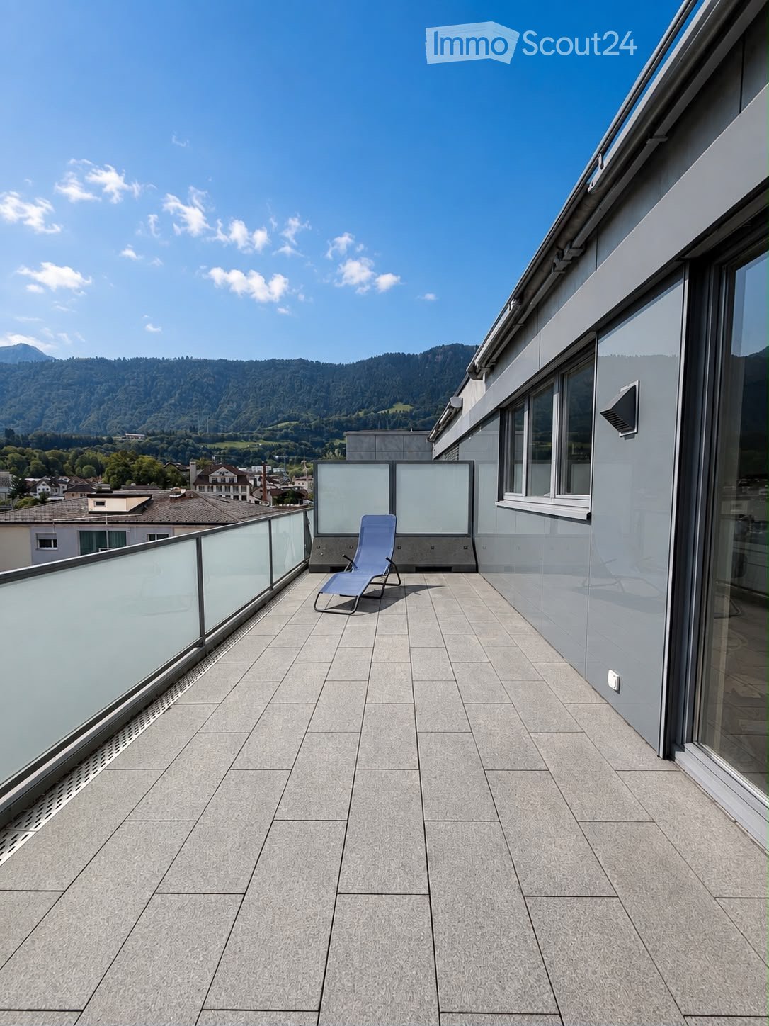 Modern rooftop terrace with grey tiled flooring, glass balustrade, and a single lounge chair, overlooking a scenic mountain landscape.