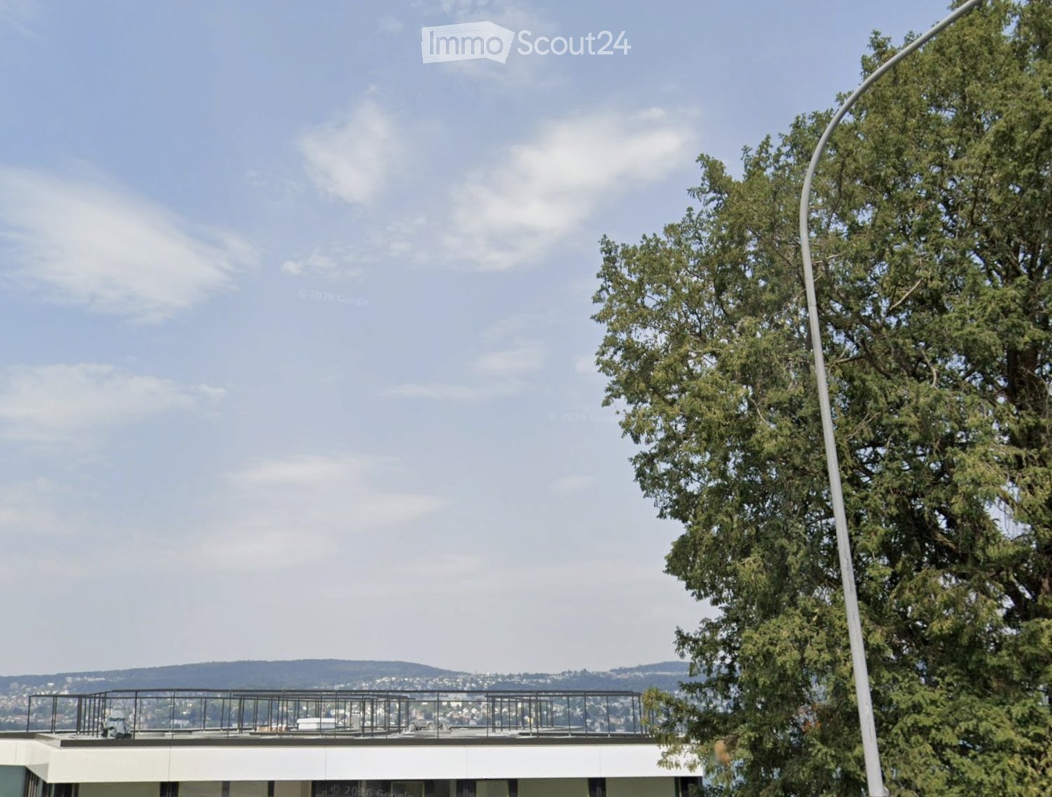 A rooftop terrace with a metal railing, overlooking a cityscape with trees and distant hills under a partly cloudy sky.