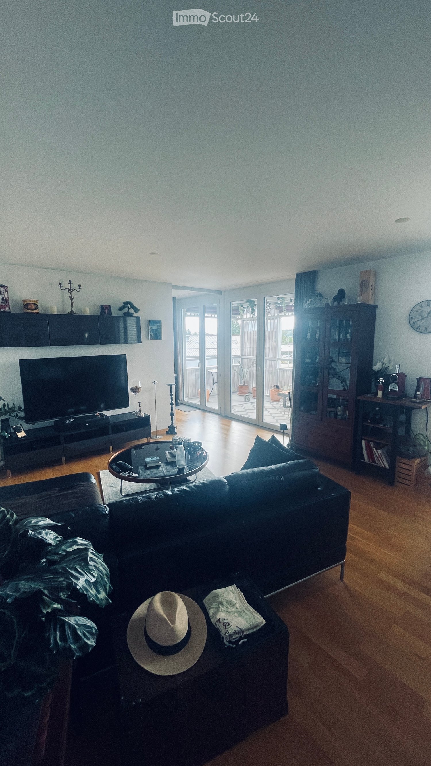 Modern living room with a black sectional sofa, coffee table, TV, and balcony access. Features wooden flooring and decorative items.