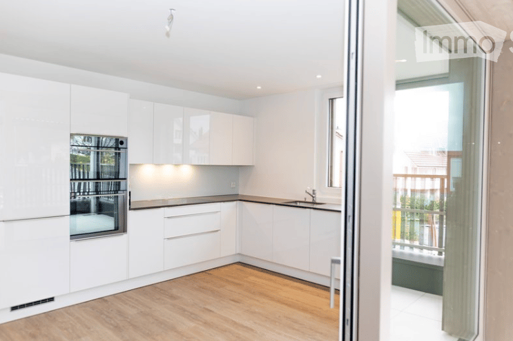 Modern, minimalist kitchen with white cabinetry, a built-in oven, and a sleek sink. The space features light wood flooring and an open sliding door leading to a balcony.
