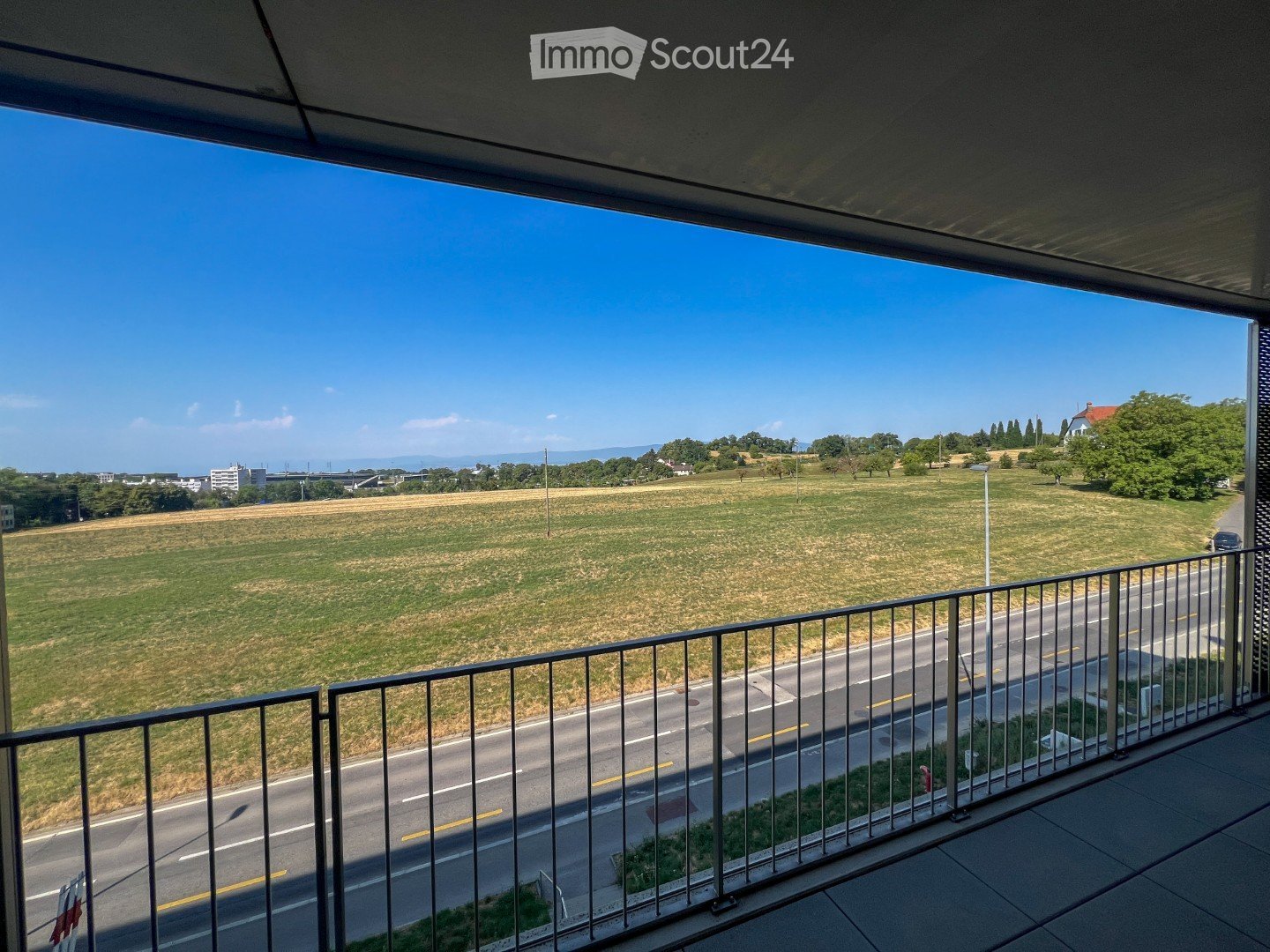 Balcony with metal railing, overlooking a road, grassy field, and distant landscape.