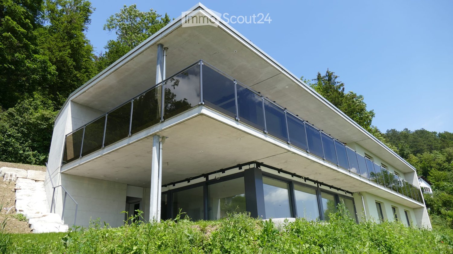 Modern two-story house with a flat roof, large glass windows, and a raised ground floor, surrounded by greenery.