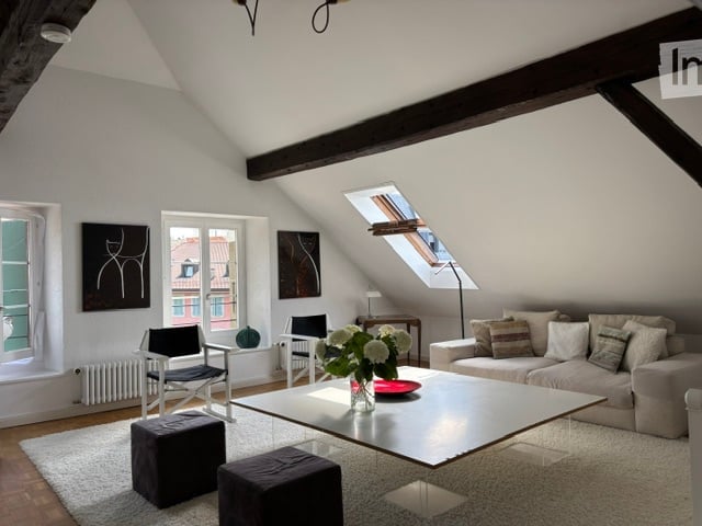 Bright living room with slanted wooden ceiling beams, large windows, and skylight. Features a grey sofa, coffee table, two black chairs, two dark grey ottomans, a small side table with a lamp, and decorative art pieces on the walls.