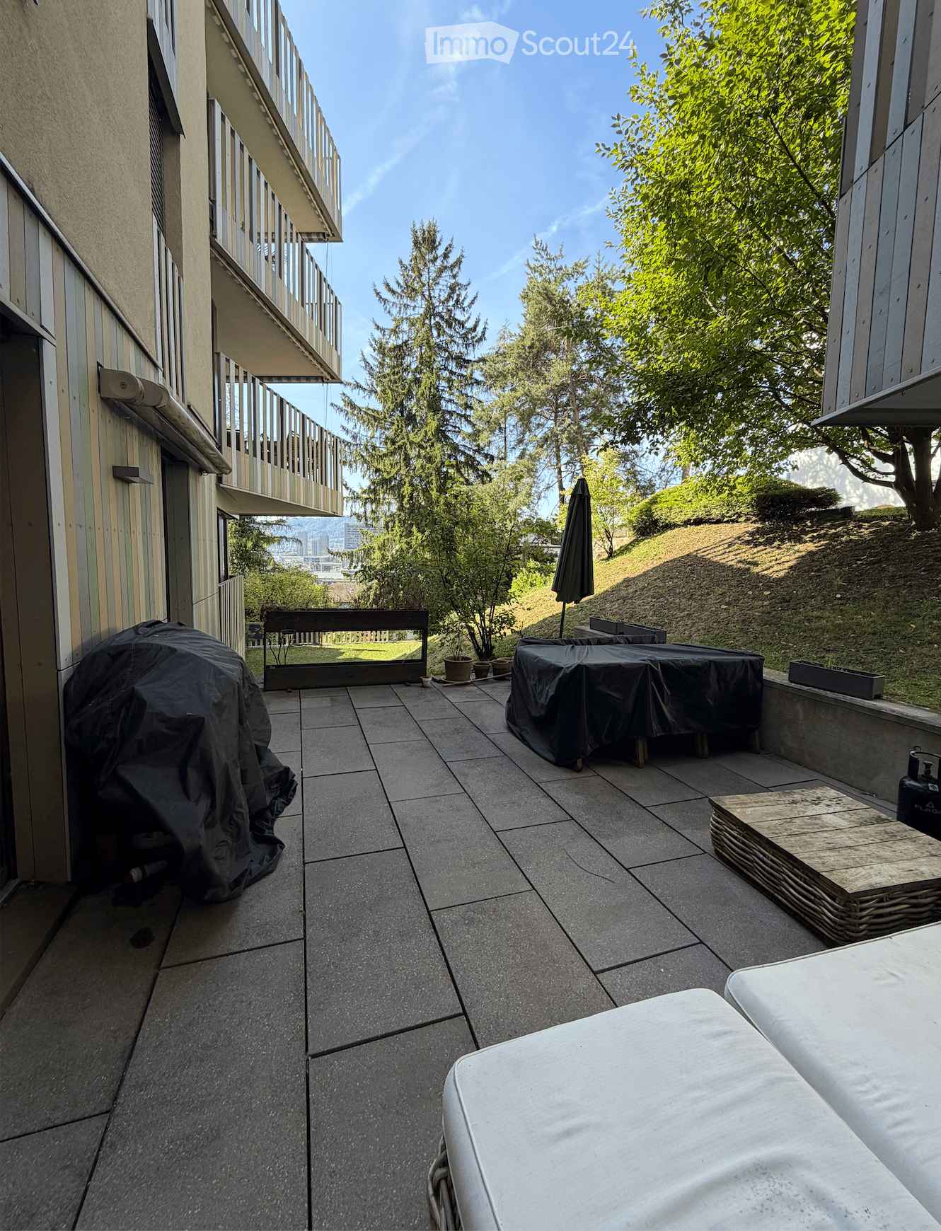 Balcony with tiled floor, covered furniture, outdoor umbrella, and view of greenery and cityscape.