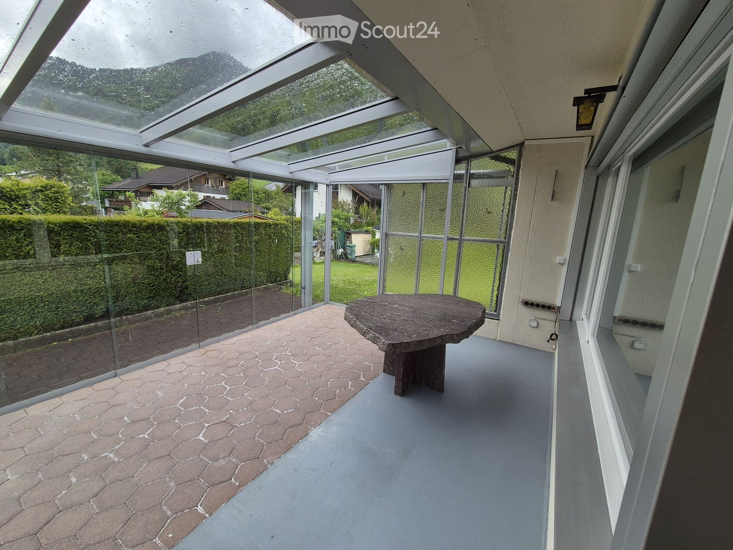Glass-enclosed balcony with a round stone table, hexagonal tiled floor, and views of a garden and surrounding houses.