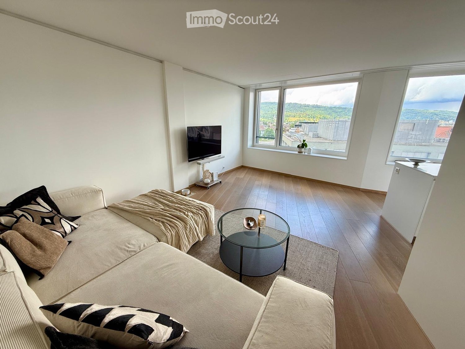 Modern living room with a large window offering a scenic view, featuring a beige sofa with patterned cushions, a round glass coffee table, and a flat-screen TV on a stand.