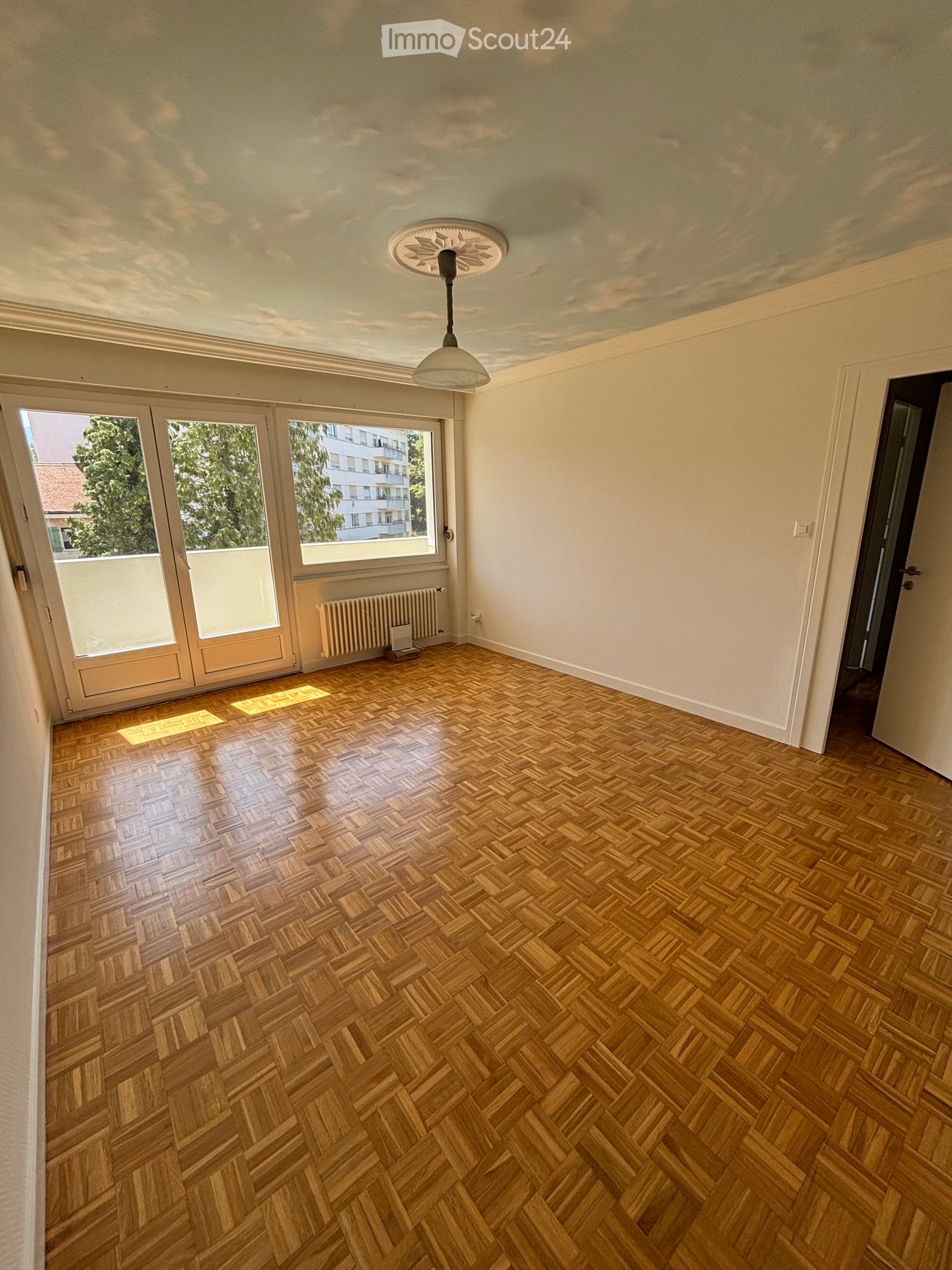 Empty room with wooden herringbone flooring, white walls, large windows with blinds, and a hanging light fixture.