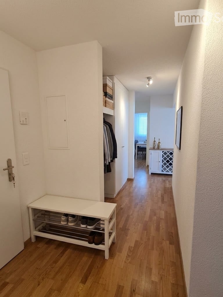 A well-lit hallway with wooden flooring, white walls, and a built-in shoe rack. A small bench with shoes is visible, and a closet with hanging clothes is on the right side. The hallway leads to another room with a dining area visible in the background.