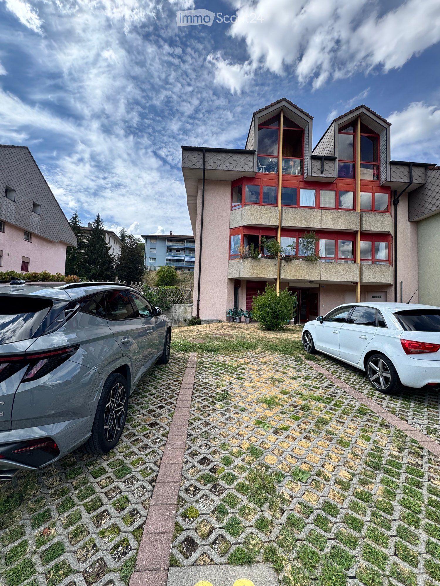 Modern two-story house with red-framed windows, balconies, and a paved driveway with two parked cars.