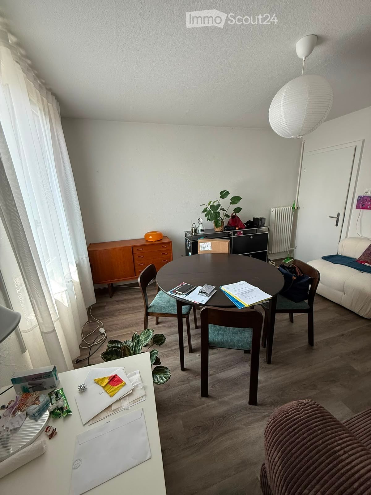 A modern, well-lit living space with a round wooden dining table, four chairs, a sofa, a wooden cabinet, a floor lamp, and a small desk with various items.