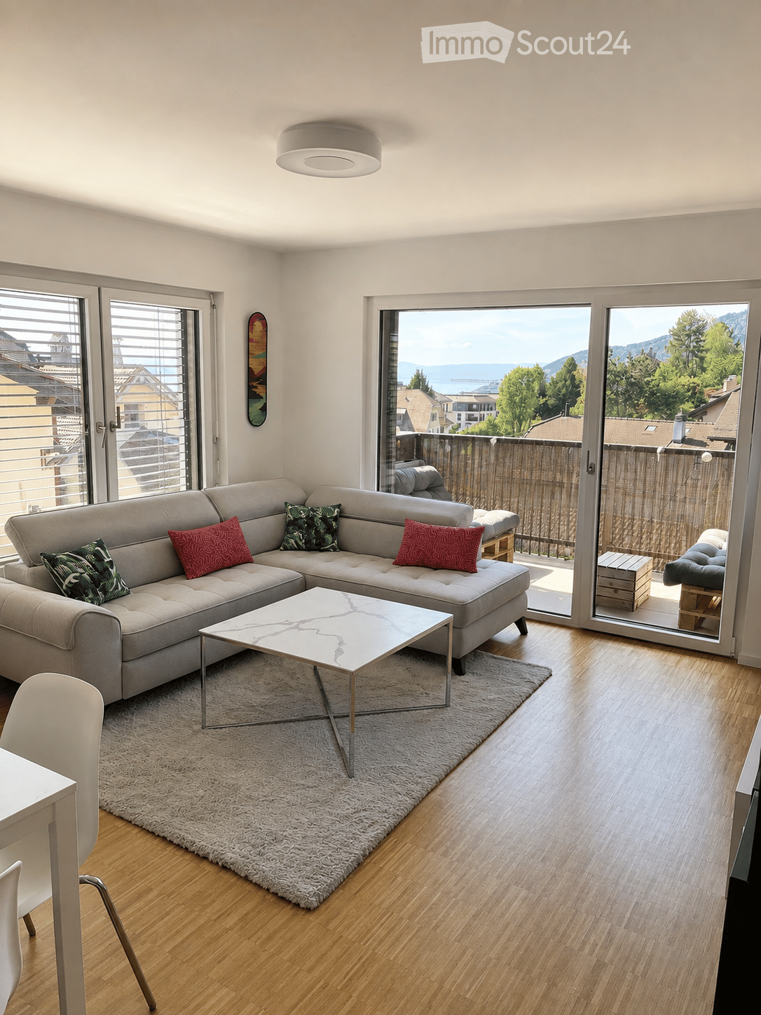 Modern living room with large windows offering a scenic view, featuring a grey sectional sofa, a white coffee table, a plush rug, and a dining area with a white table and chairs.