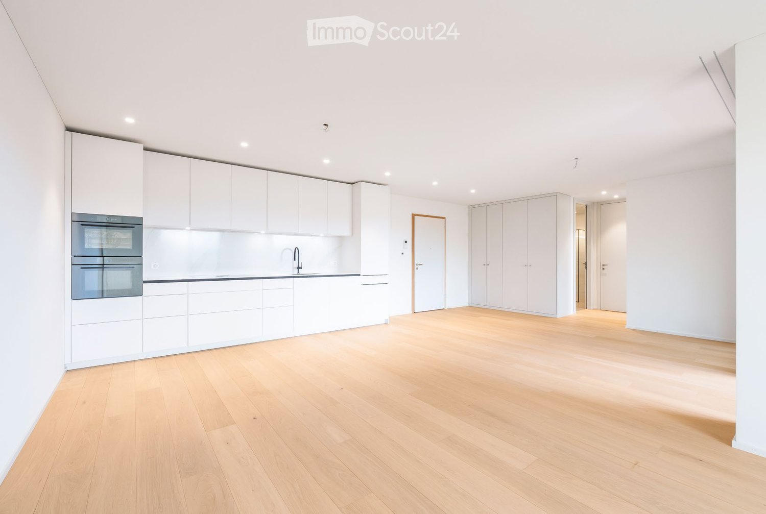 Bright, open-plan space with light wooden flooring, white cabinetry, and built-in appliances including a double oven and sink.