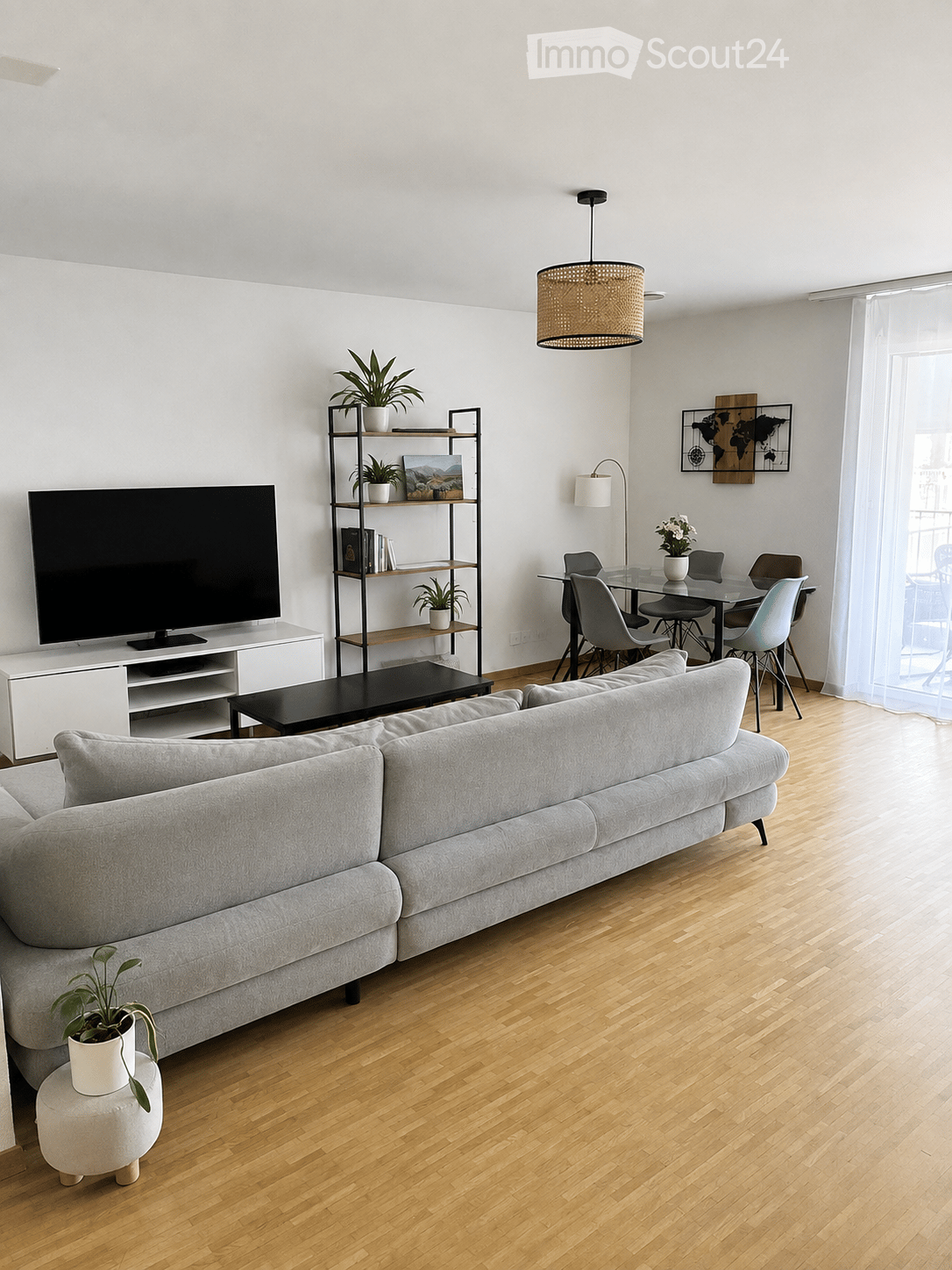 Modern living room with light wooden flooring, grey sectional sofa, coffee table, TV, shelving unit with plants and books, dining area with glass table and chairs, and a hanging woven pendant light.