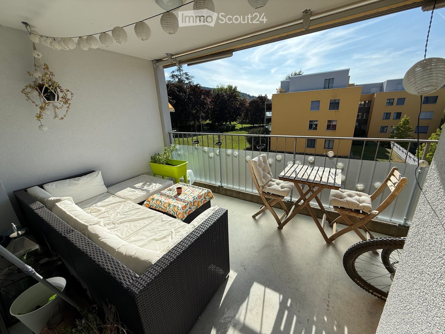 Balcony with a white sofa, small table, two wooden chairs, and a bicycle wheel. The balcony has a glass railing and a view of a park and buildings.