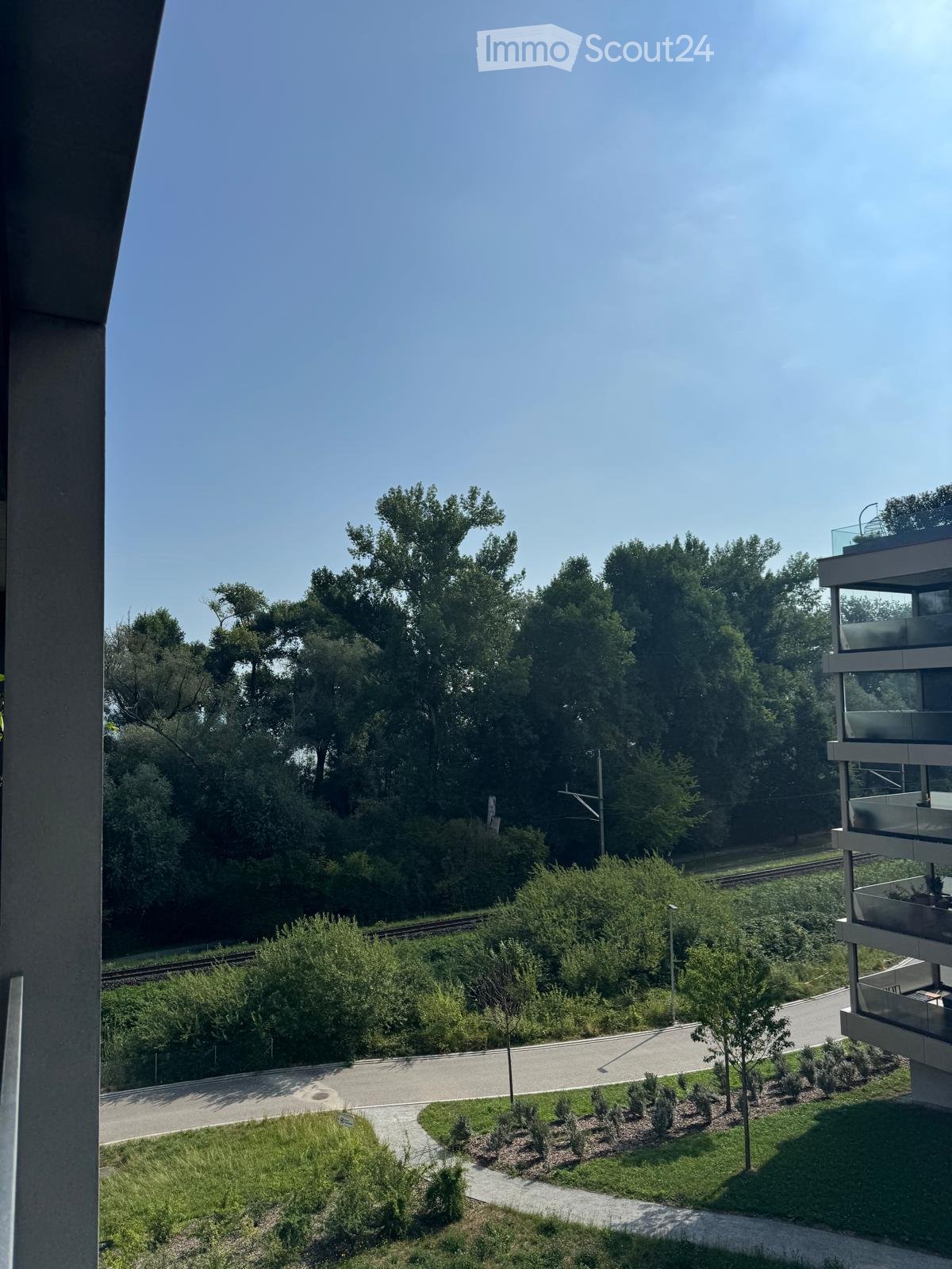 Balcony view overlooking a landscaped area with trees, a pathway, and a modern building with glass railings.