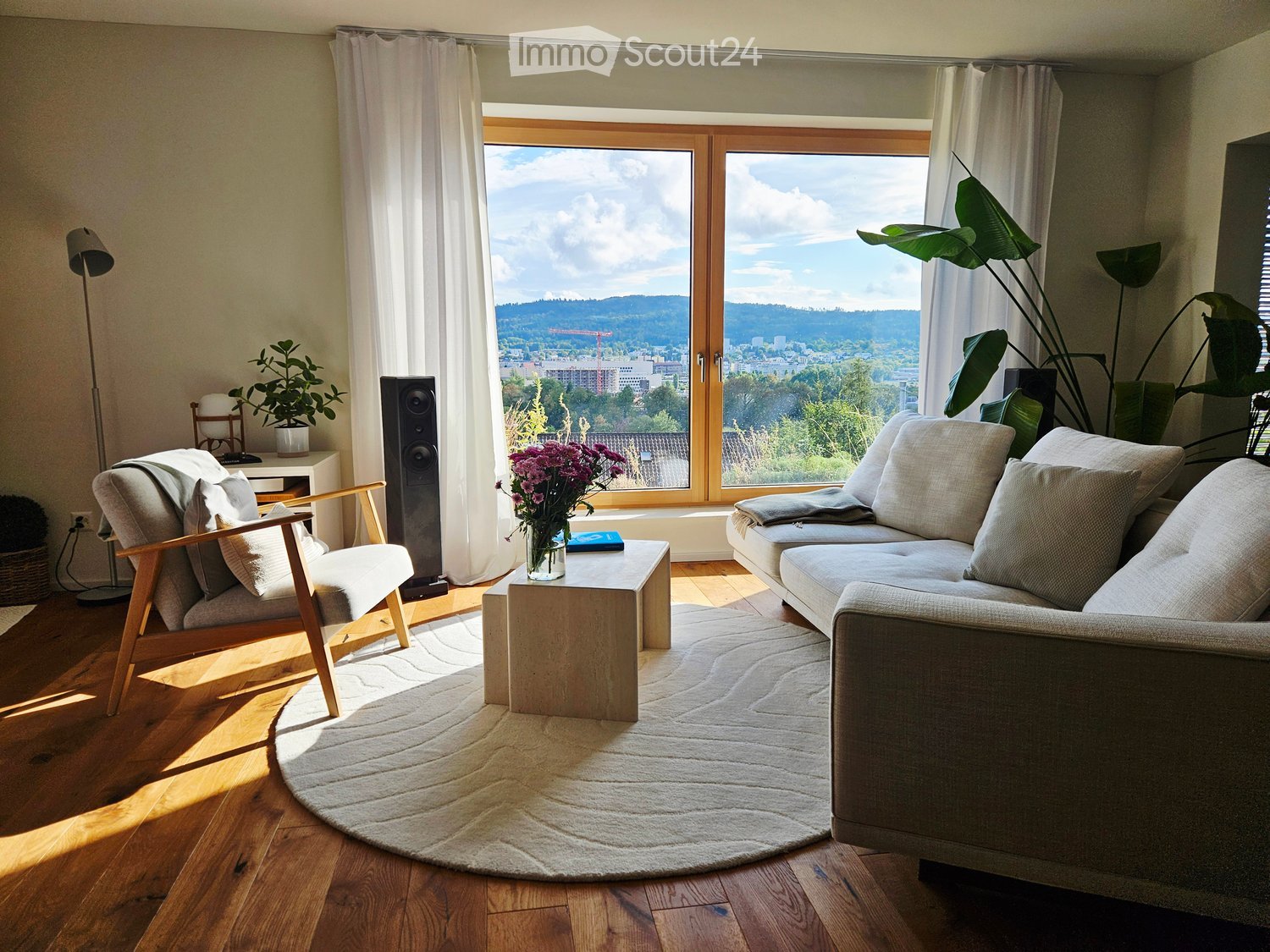 Bright living room with large windows offering a scenic view, featuring a sofa, armchair, coffee table, floor lamp, and decorative plants.