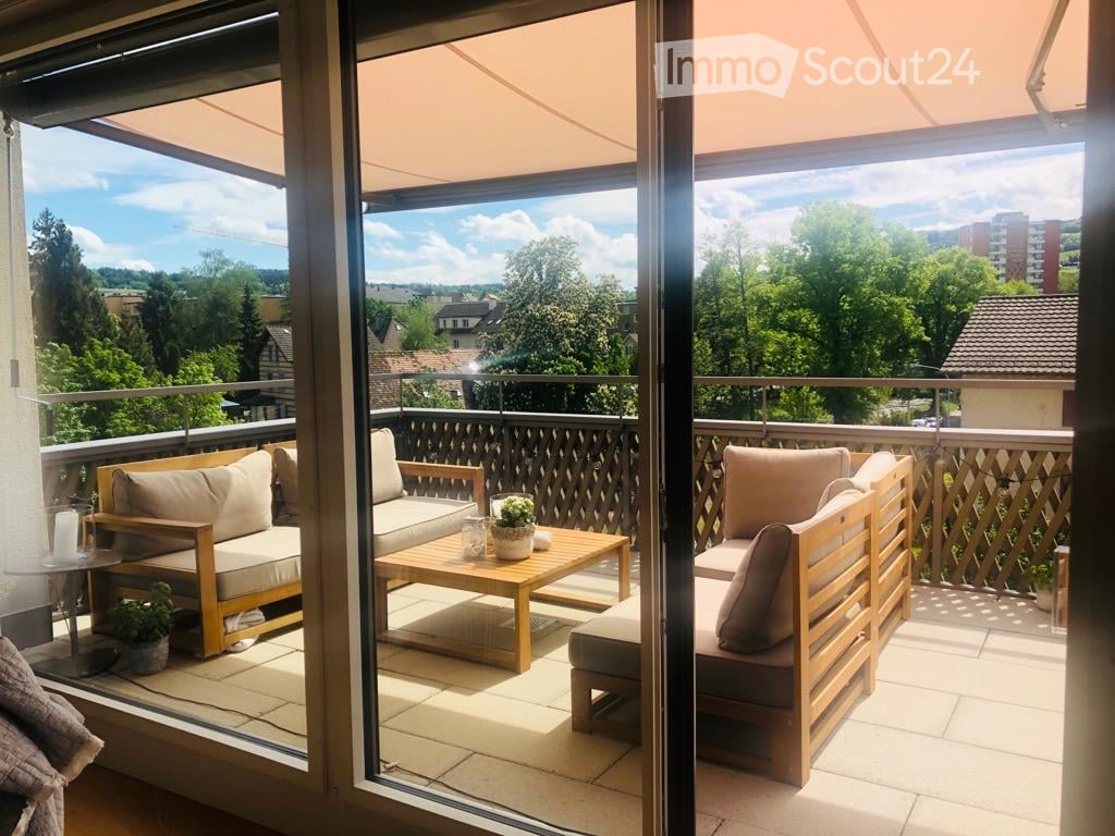 Balcony with wooden furniture, including a sofa, coffee table, and chairs, with a scenic view of trees and buildings.