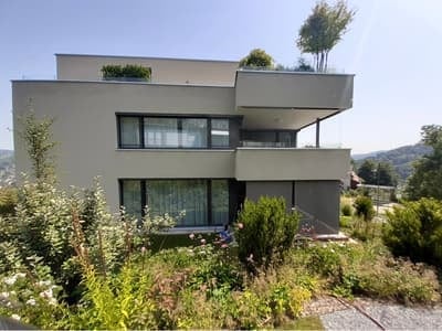Modern two-story house with flat roof, large windows, and balcony on the upper floor. The house is surrounded by a well-maintained garden with various plants and shrubs.