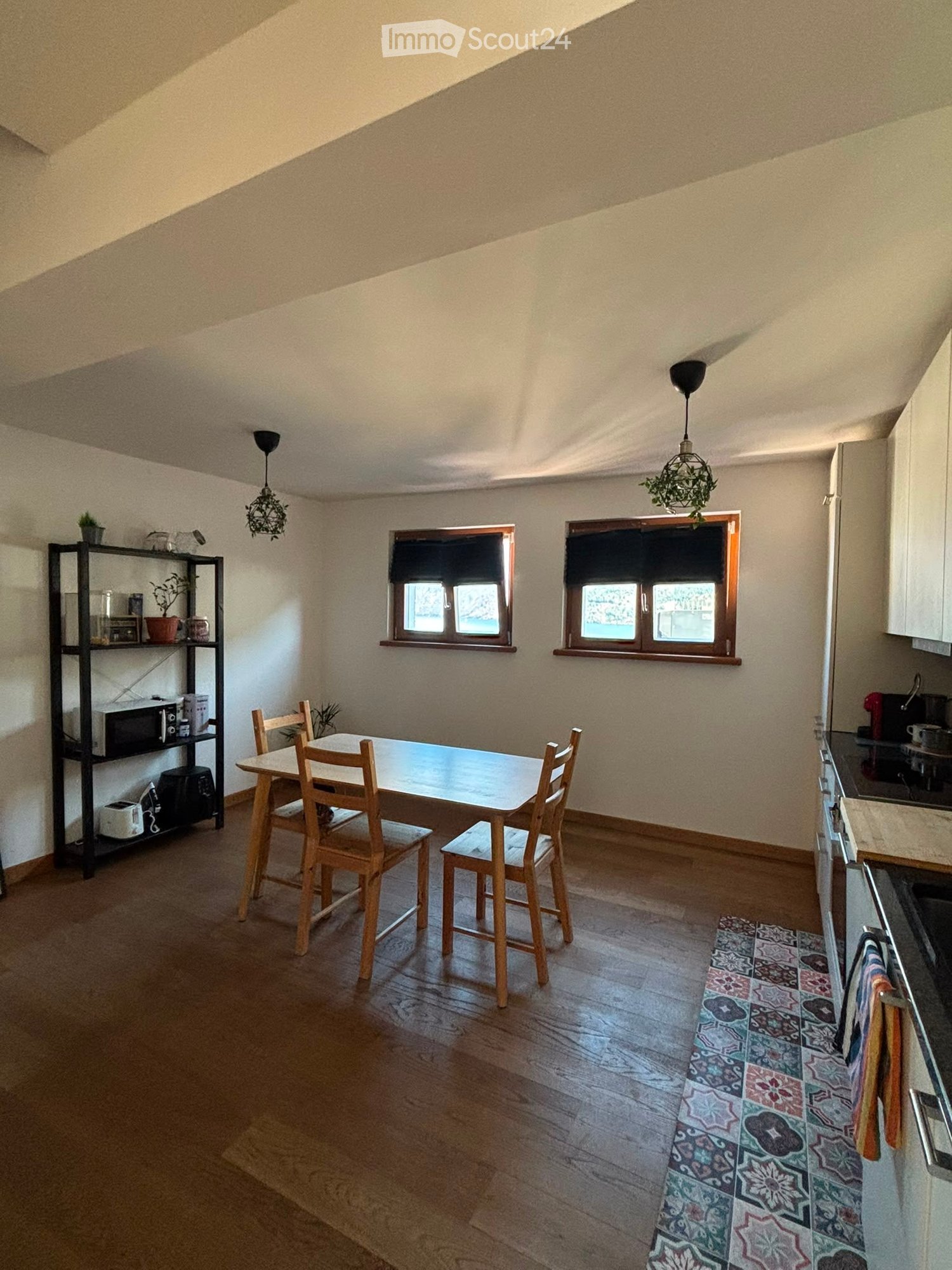 Dining area with a wooden table, four chairs, and a stool, adjacent to a small kitchen area with a counter, sink, and pendant lights. A shelf with a microwave and toaster is visible, along with a colorful rug on the floor.