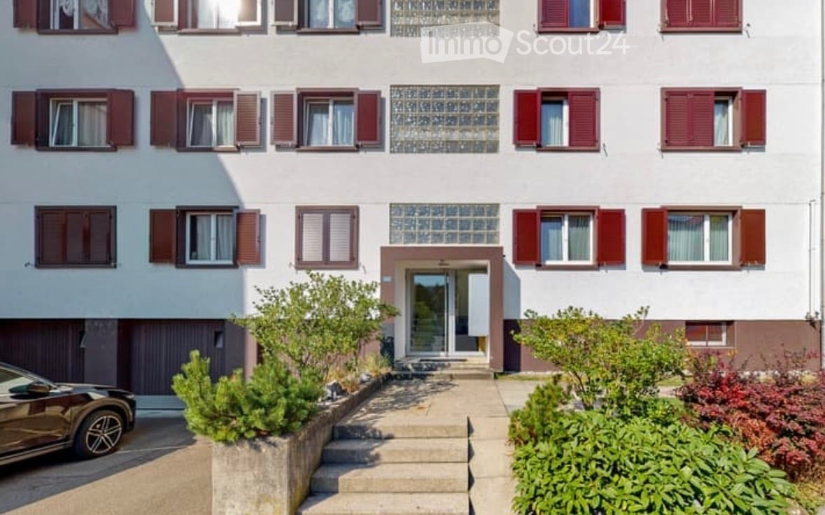 A two-story residential building with white walls, red shutters, and multiple windows. The entrance features a small staircase leading to a glass door, surrounded by small shrubs and plants.