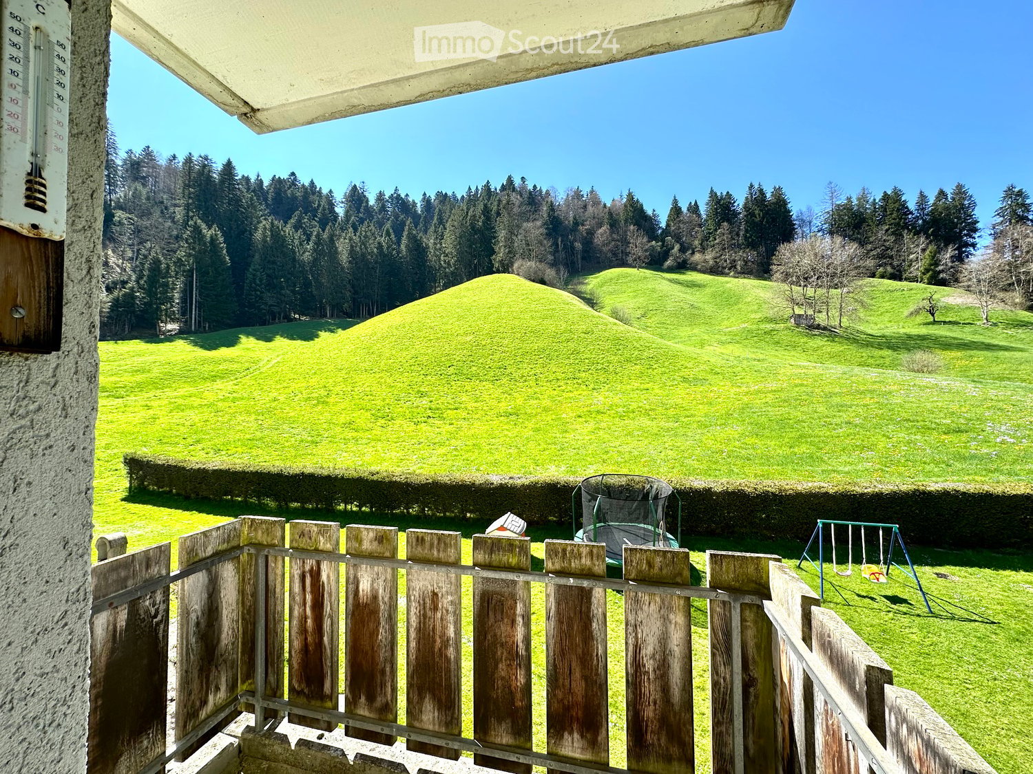 A fenced garden area with a swing set and a bouncy house, overlooking a green, hilly landscape with trees.
