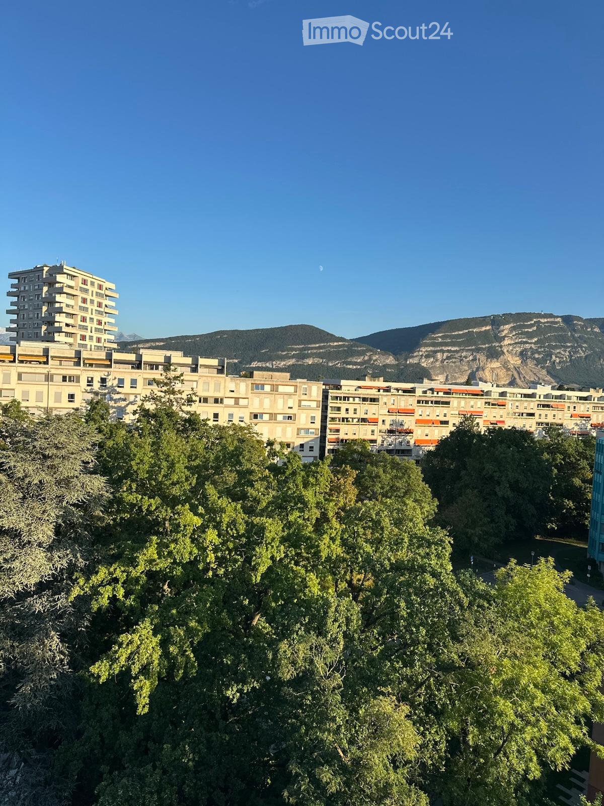 A view of a cityscape with multiple residential buildings, surrounded by lush green trees, with a mountain range in the background under a clear blue sky.