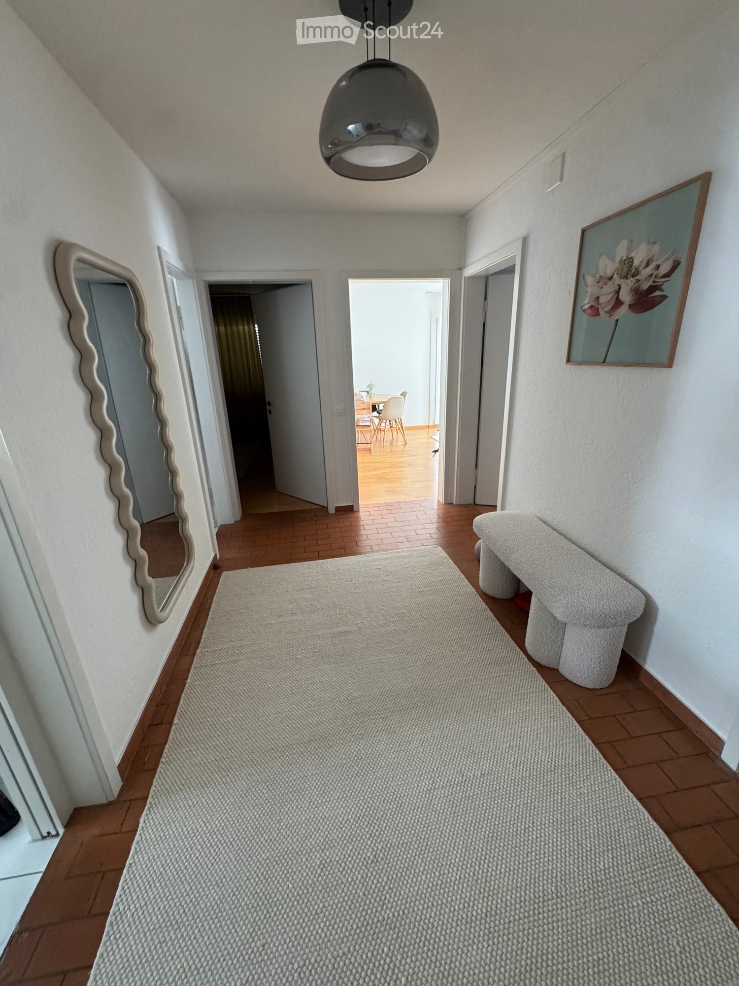 Hallway with wooden flooring, a large mirror on the left, a white bench on the right, and a painting on the wall. The hallway leads to a dining area with a table and chairs.