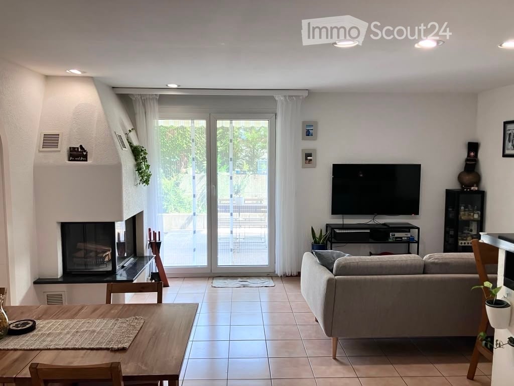 Modern living room with a grey sofa, coffee table, flat-screen TV, fireplace, dining table, and potted plants. Large sliding glass doors lead to a patio with outdoor seating.