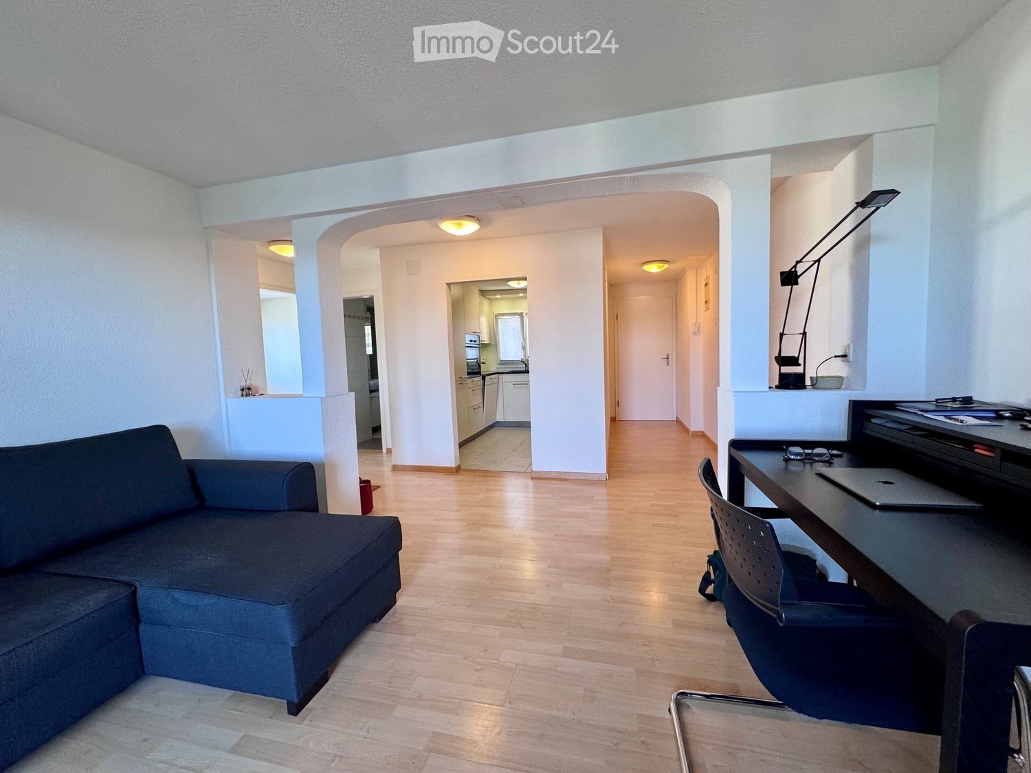 Modern living room with a dark blue sectional sofa, a wooden floor, a desk with a laptop, and a view into a small kitchen area with white cabinets and an oven.