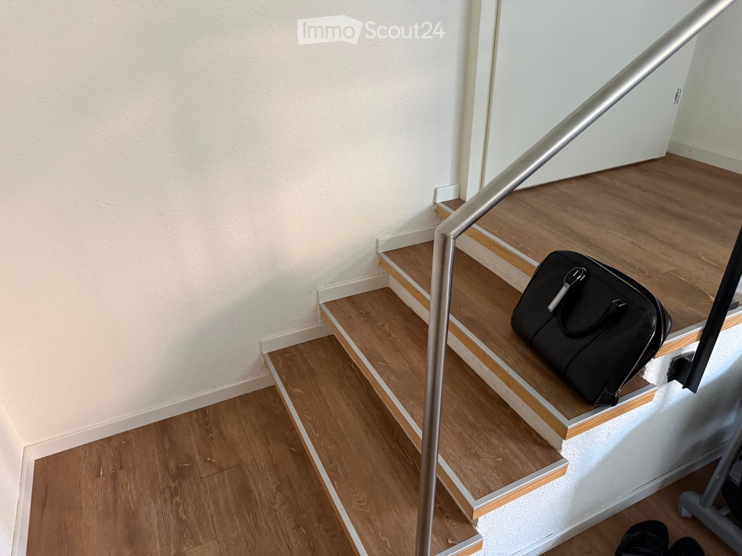 Wooden staircase with a metal handrail, a black bag placed on one of the steps, and a white wall in the background.