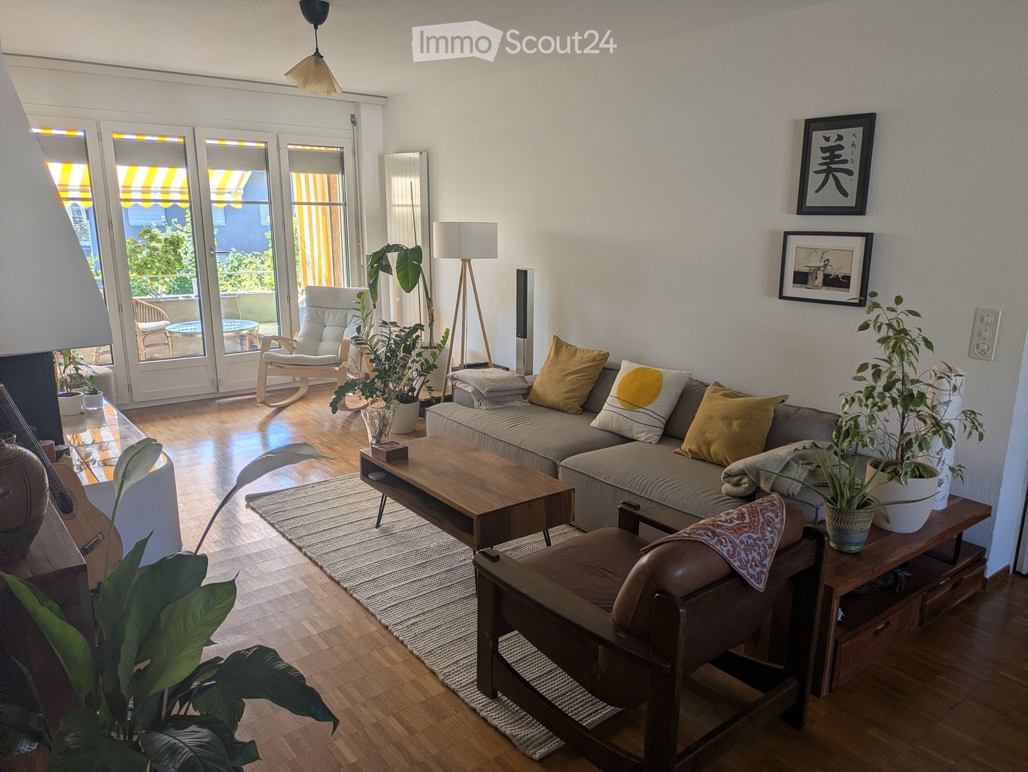 Modern living room with a grey sofa, wooden coffee table, armchair, floor lamp, and potted plants. Large windows and sliding doors lead to a balcony.