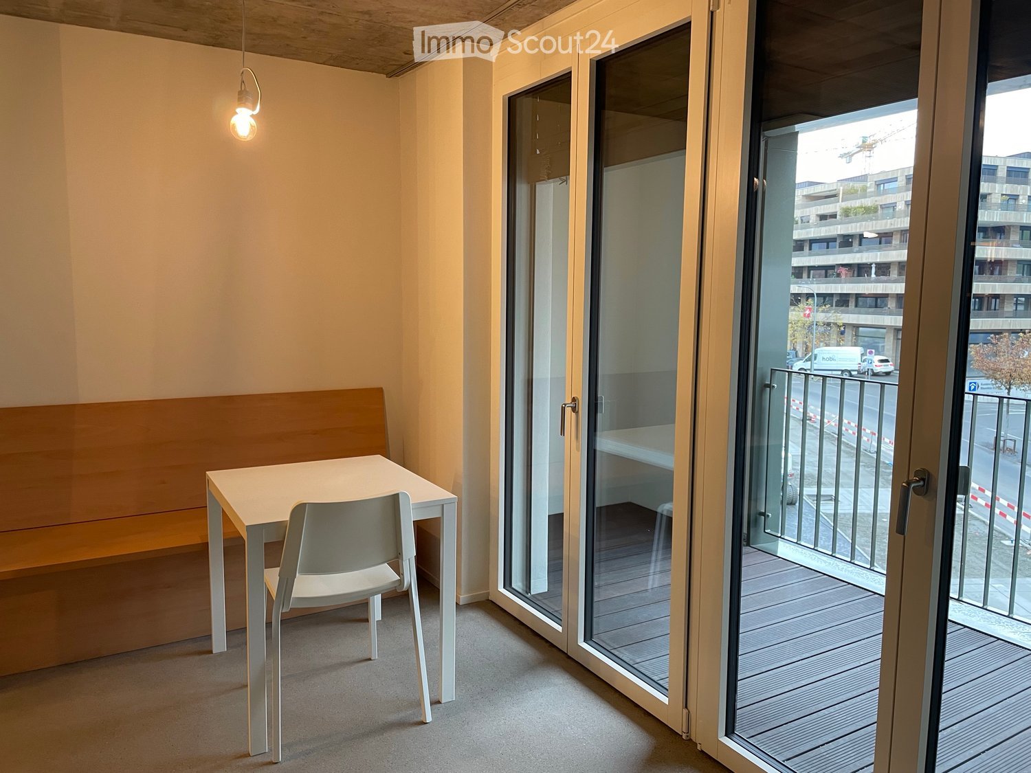 A small, modern room with a white table and chair, a built-in bench, and large glass doors leading to a balcony.
