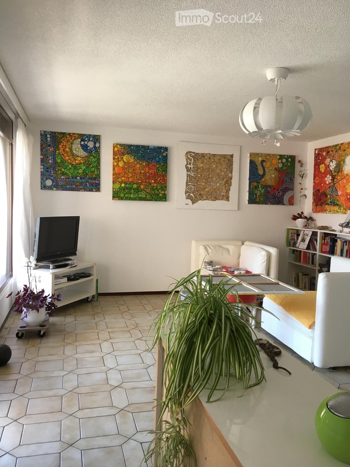 A modern living room with white walls, hexagonal tiled flooring, and large windows. Features include a white sofa, a glass coffee table, a television on a white stand, a bookshelf, and several colorful abstract paintings on the walls.