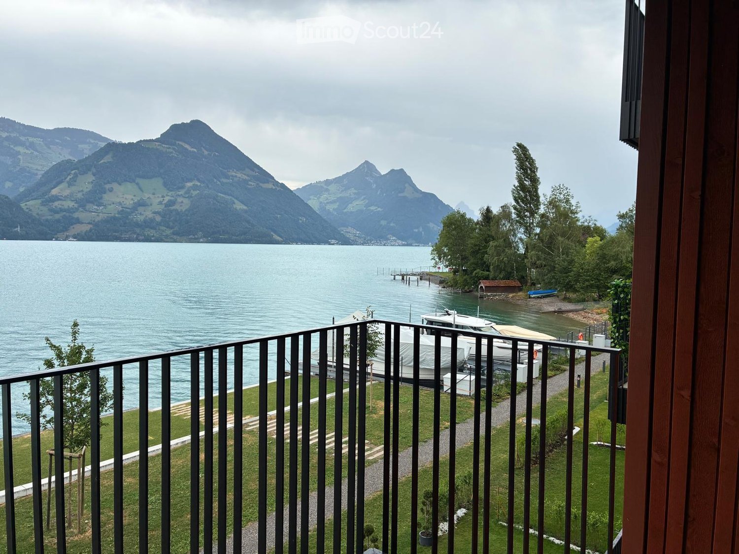 Balcony overlooking a lake with mountains in the background, featuring a metal railing and a view of boats and a dock.