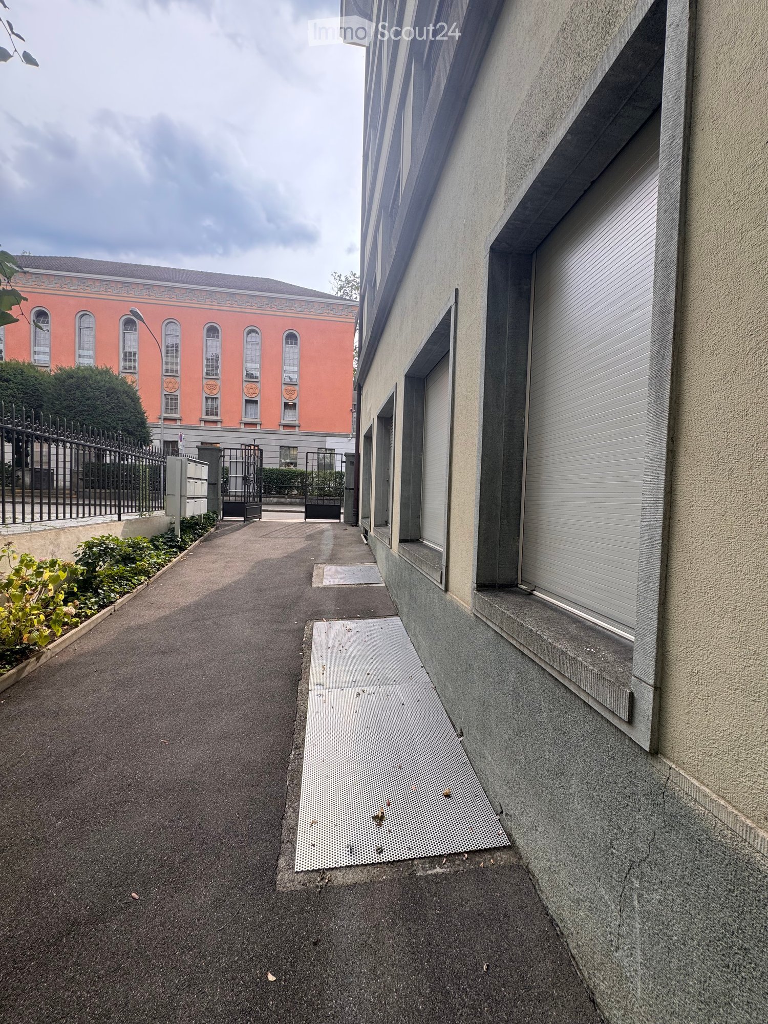 Narrow paved pathway between buildings, with metal grates, small garden patches, and a gated entrance leading to a larger red building.