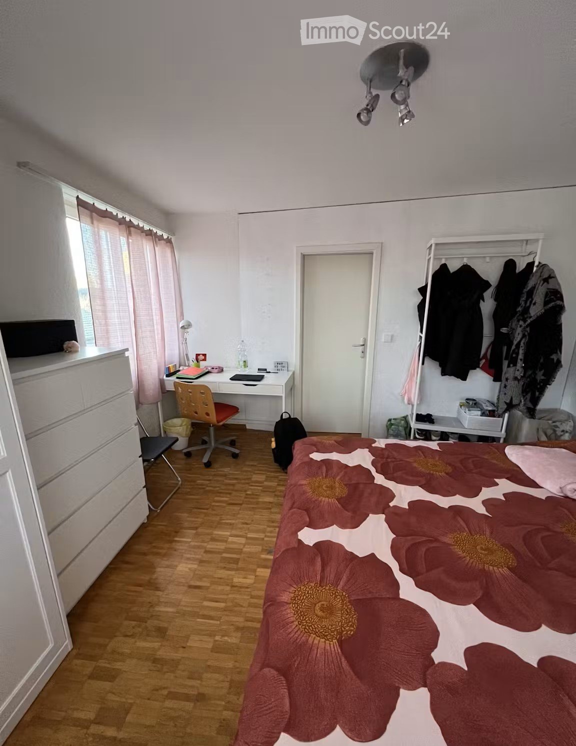 A well-lit bedroom with wooden flooring, a large bed with a floral duvet, a desk with a chair, a wardrobe with hanging clothes, and a window with curtains.