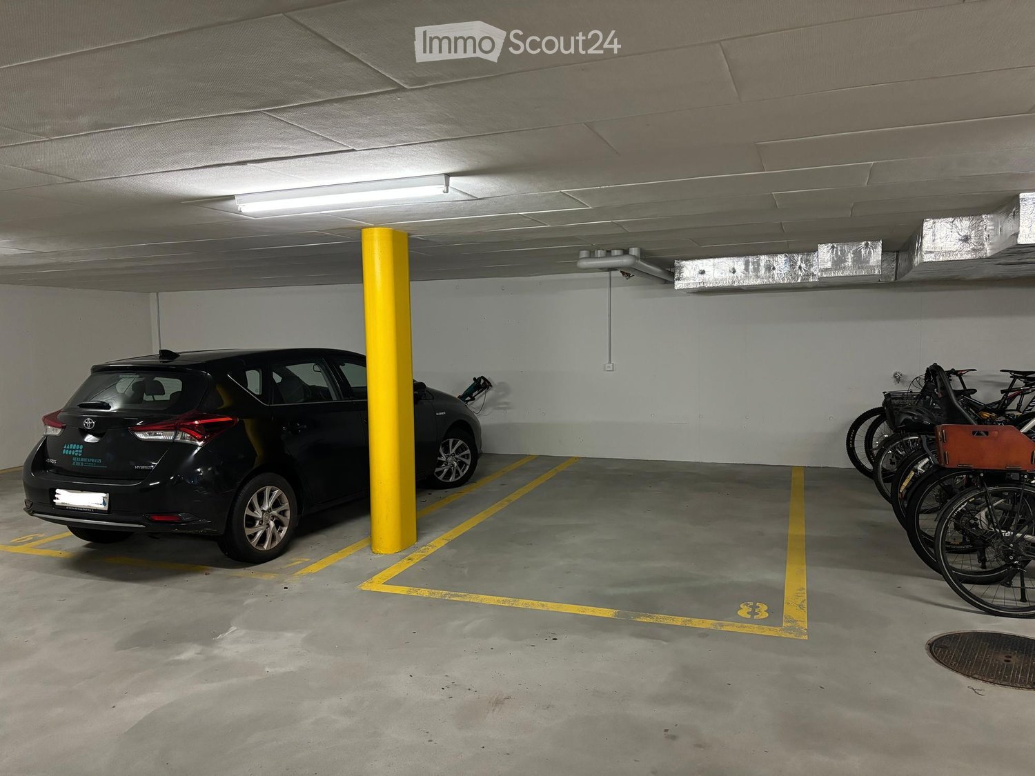 Underground parking area with a parked car and several bicycles, marked parking spaces, and a yellow support pillar.