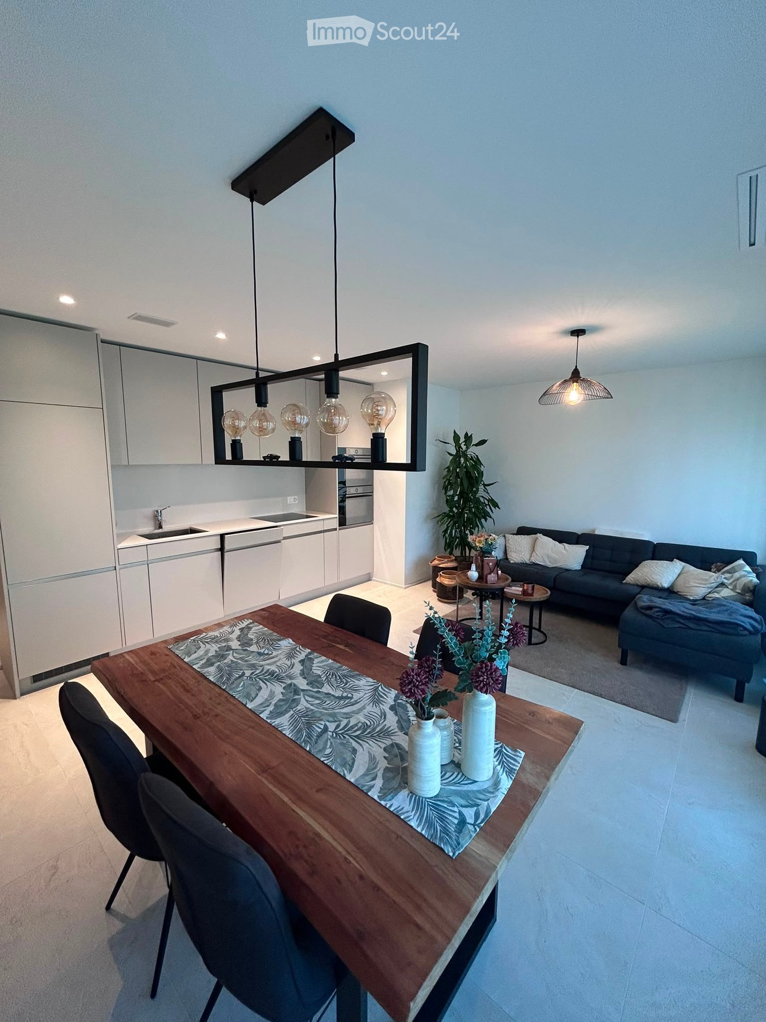 Modern open-plan living and dining area with a dark grey sofa, wooden dining table, pendant lights, and a sleek white kitchen with cabinets and appliances.