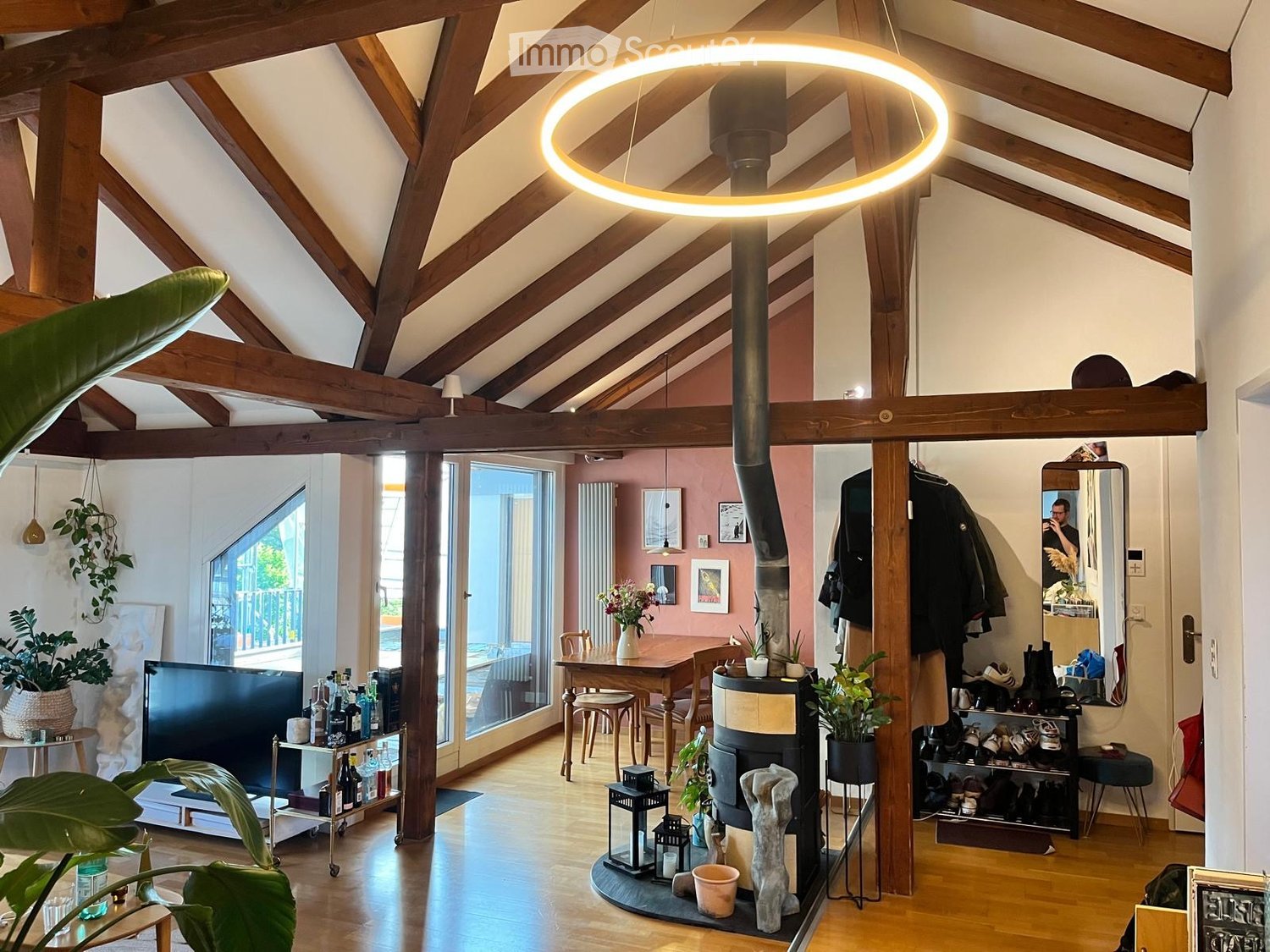 Open-plan living and dining area with wooden beams, exposed wooden floor, large windows, fireplace, dining table, sofa, TV, and various decorative items.