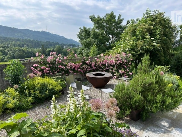 Well-maintained garden with a variety of flowering plants, shrubs, and trees. Features include a stone wall, a large decorative bowl, and a small seating area with chairs and a table.