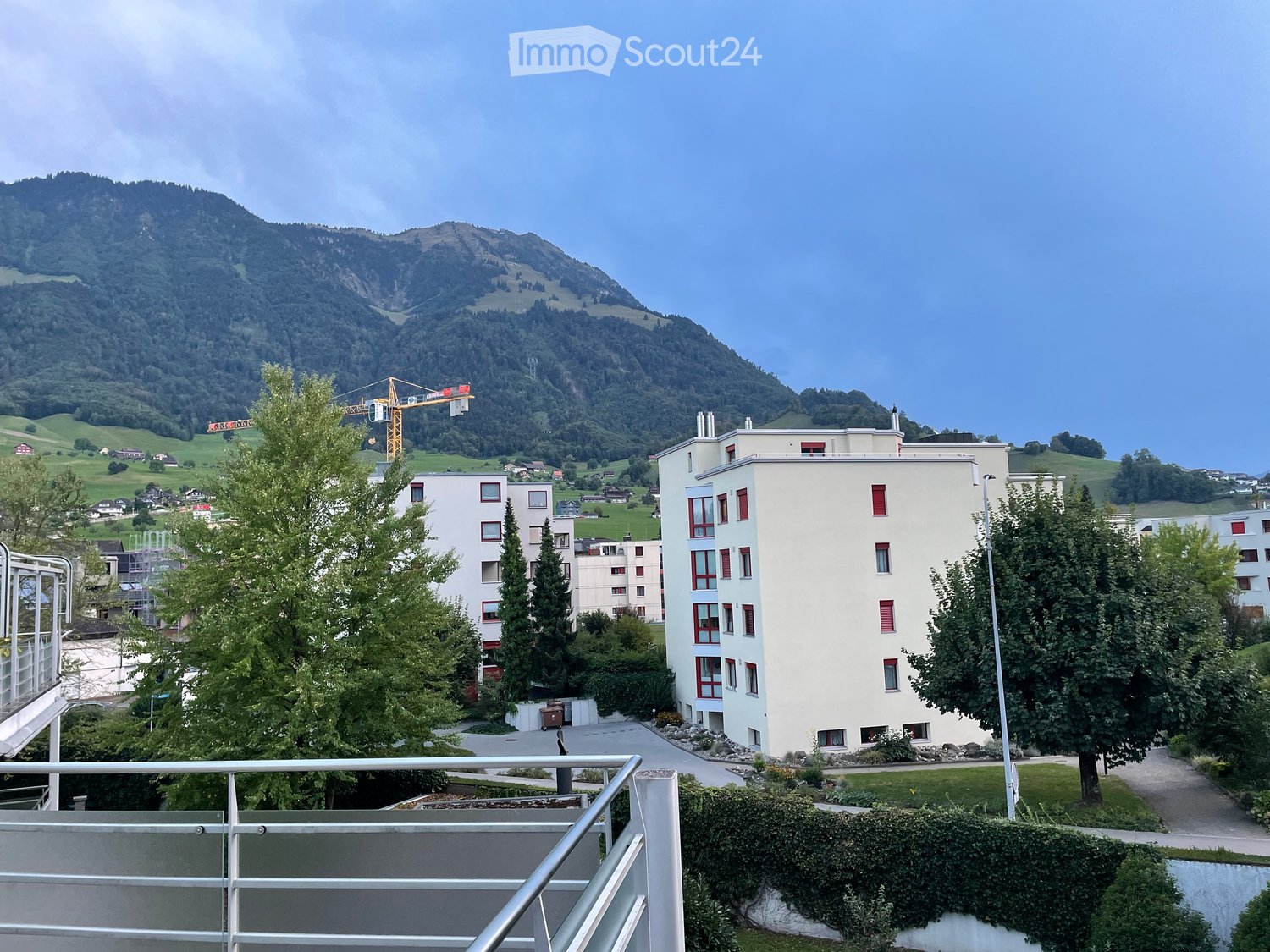 View of a modern residential building with red-framed windows, surrounded by trees and a mountainous landscape in the background.