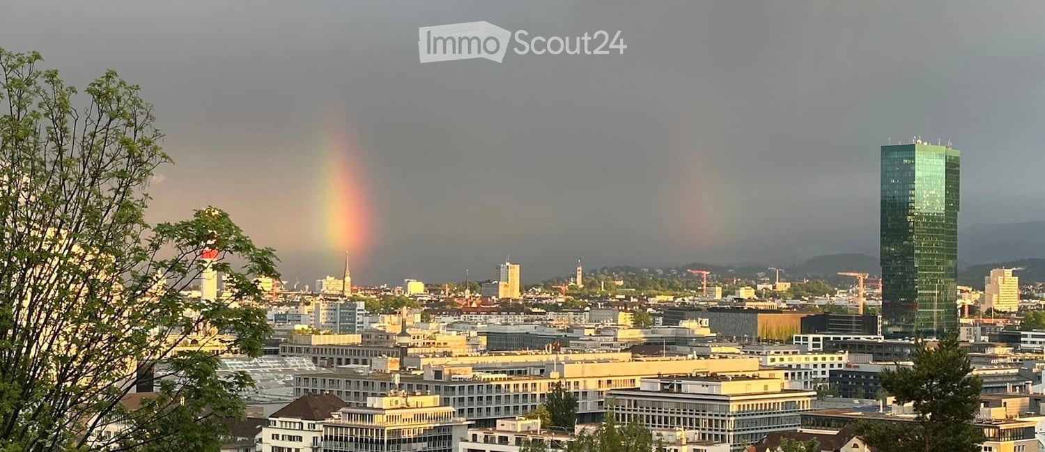 A panoramic view of a cityscape with a rainbow in the sky, featuring a mix of modern and older buildings, trees, and distant mountains.