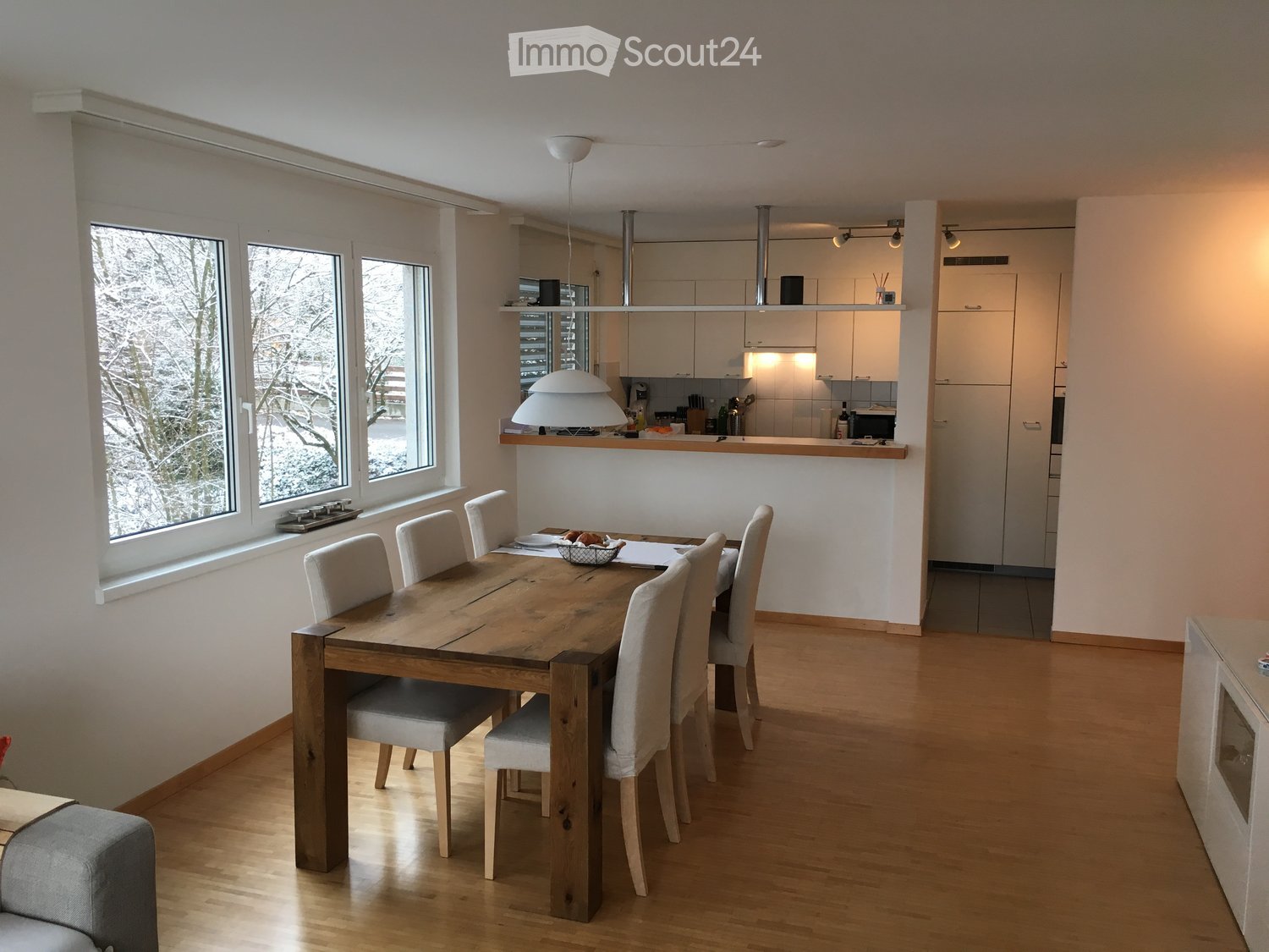 Open-plan dining and kitchen area with wooden flooring, large windows, a wooden dining table with six chairs, and a modern kitchen with white cabinets and stainless steel appliances.