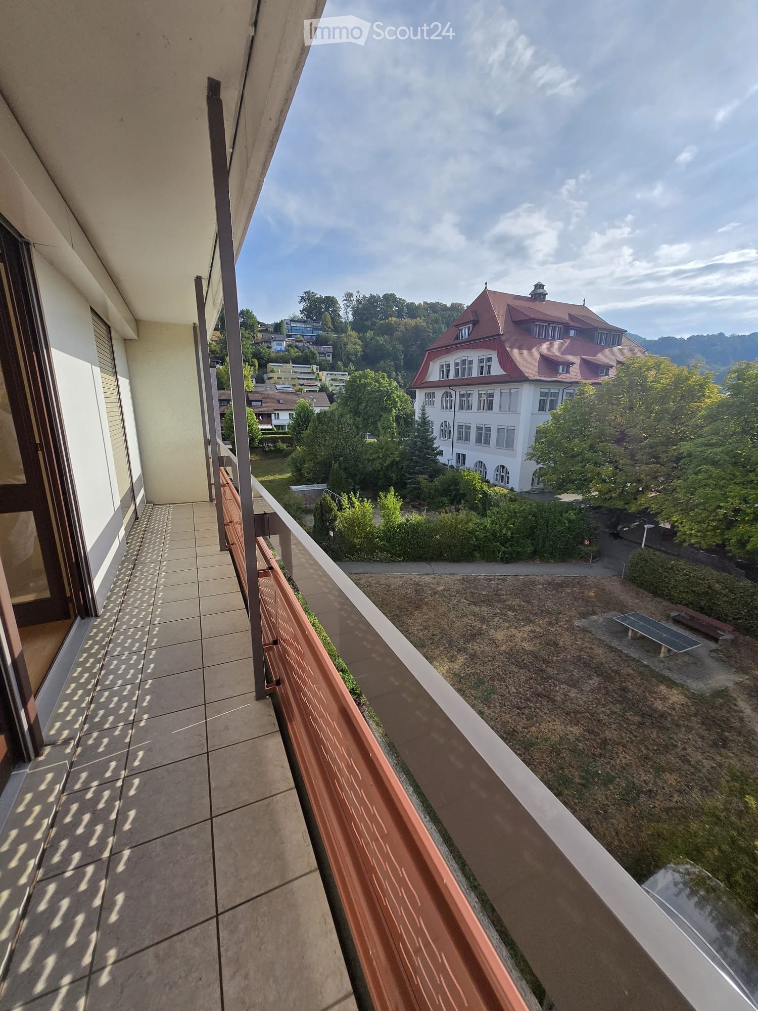 Balcony with tiled floor and wooden railing, overlooking a park area with benches and a large building in the background.
